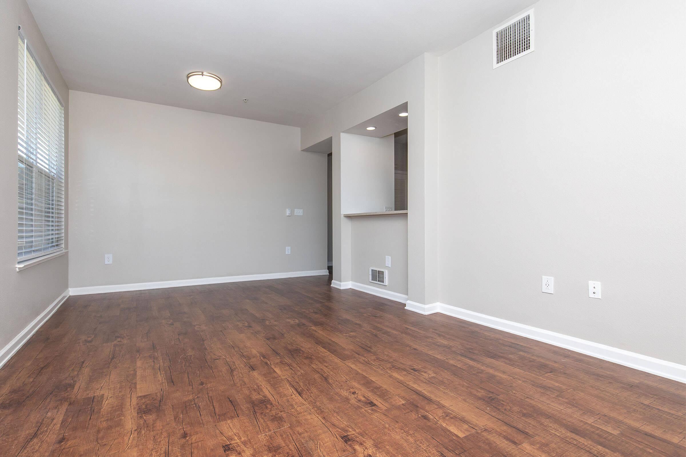 A spacious, light-colored room with hardwood flooring, featuring a single ceiling light fixture, a large window with blinds on one wall, and an opening to a kitchen area in the background. The walls are painted in a neutral tone, creating a bright and open atmosphere.