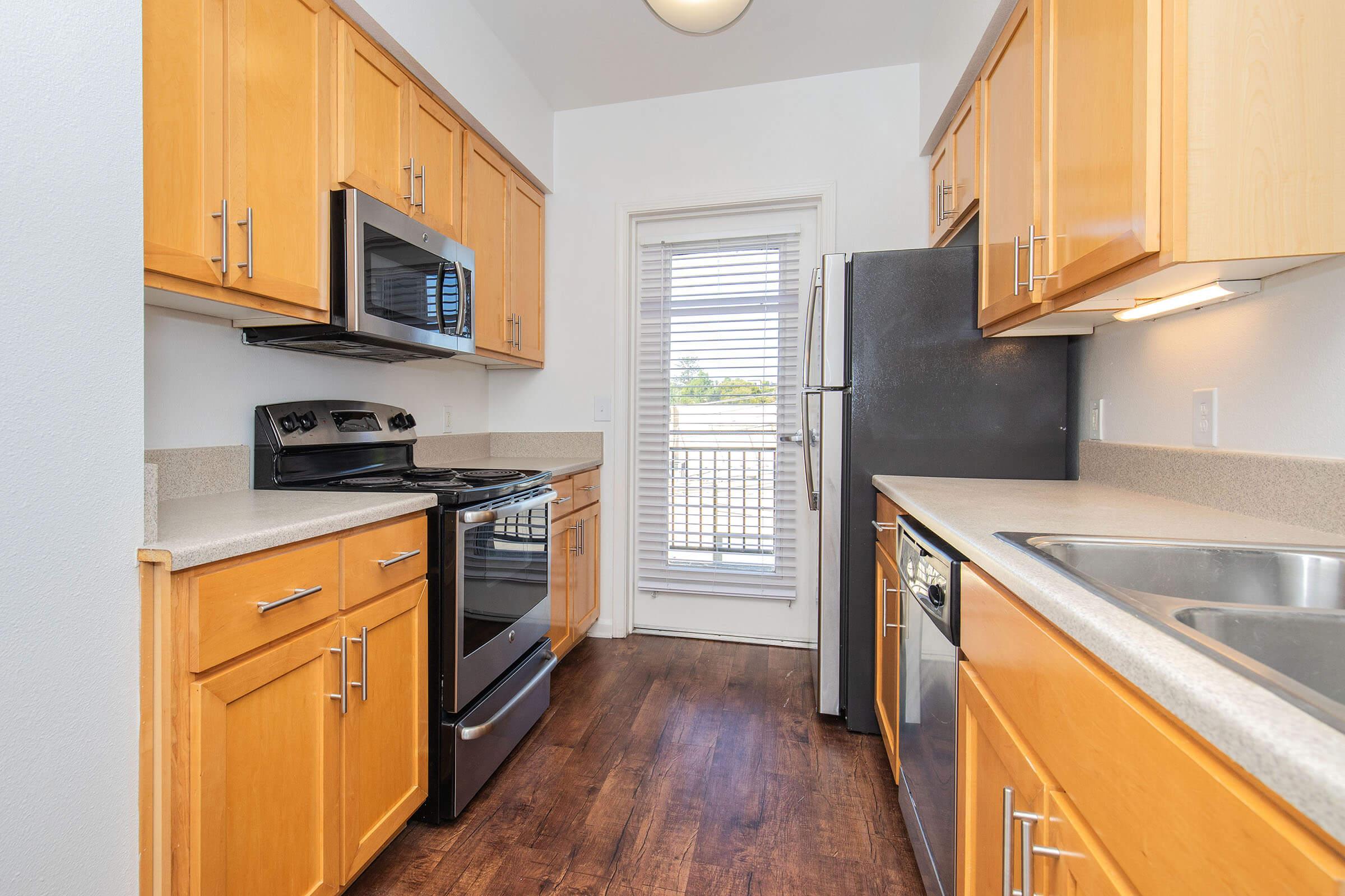 A modern kitchen featuring wooden cabinetry, stainless steel appliances including a microwave, oven, and refrigerator. A double sink sits beside the dishwasher, and a window provides natural light, with a view of a balcony. The floor is a warm wood finish, enhancing the inviting atmosphere.