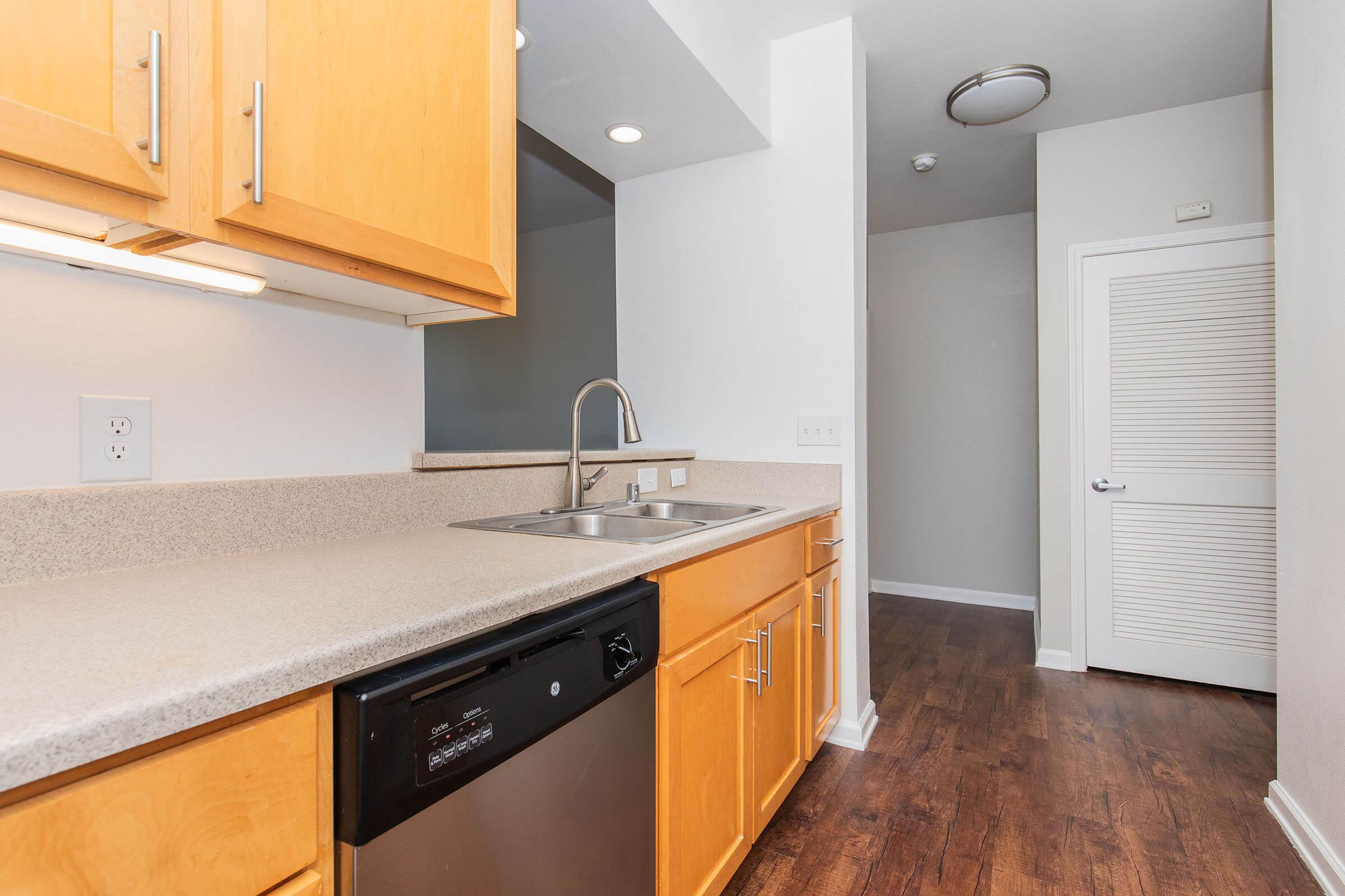 A modern kitchen featuring light wood cabinetry, a stainless steel sink, and a dishwasher. The countertop is a light stone color, and the kitchen connects to a hallway with a closed door and a light gray wall. The flooring is a medium hardwood, providing a warm and inviting atmosphere.