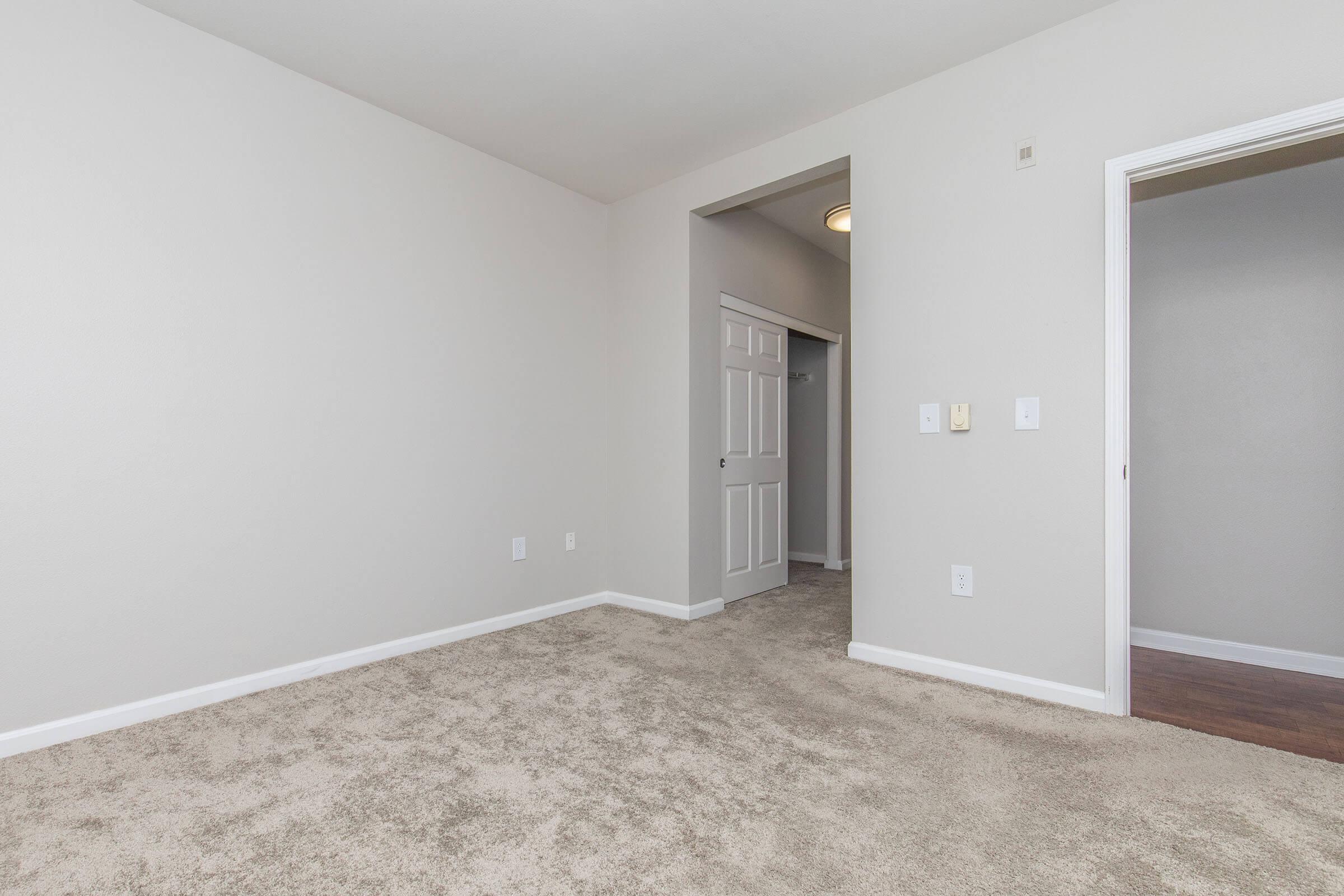 Empty room with light beige walls and a carpeted floor. There's a doorway on the right leading to a closet and another door in the background. The space is well-lit with a ceiling light fixture, creating a spacious and neutral atmosphere, ideal for personal customization.