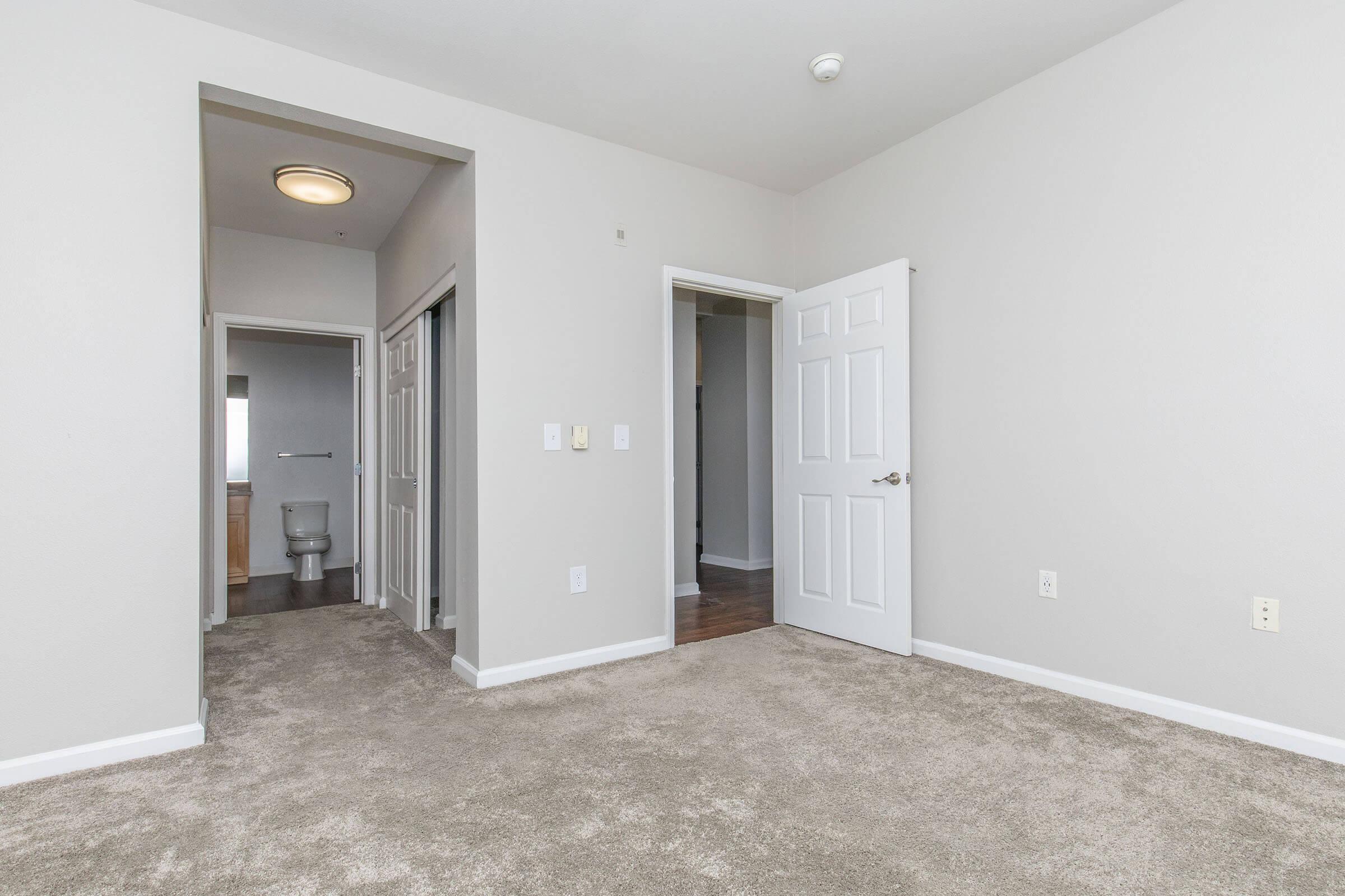 Interior view of a light-colored room with beige carpet. Two doors lead to adjacent areas, one with a visible bathroom. The walls are painted in a neutral shade, and the ceiling features a round light fixture. A small hallway is visible in the background, creating an open and spacious feel.