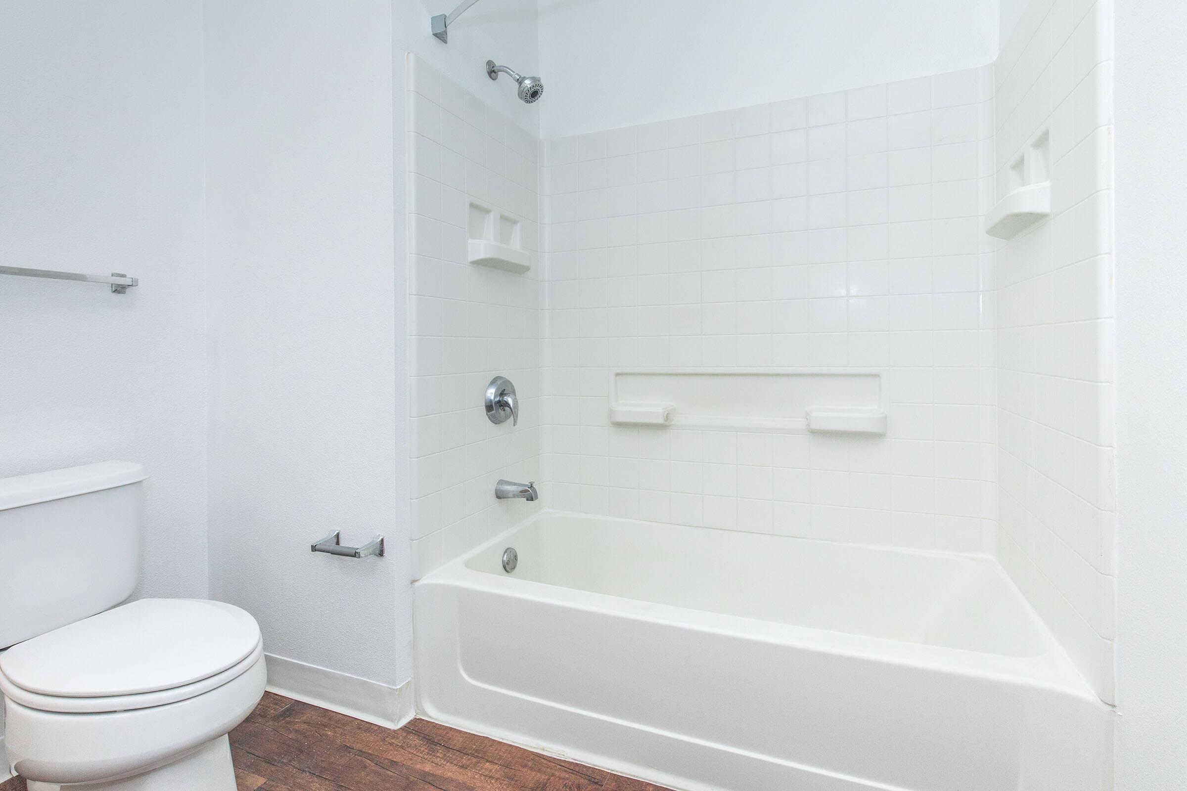 A clean, well-lit bathroom featuring a white bathtub with a showerhead above, grab bars on the wall, and a white toilet nearby. The walls are tiled in white, and the floor displays a wood-like finish, creating a modern and functional space.