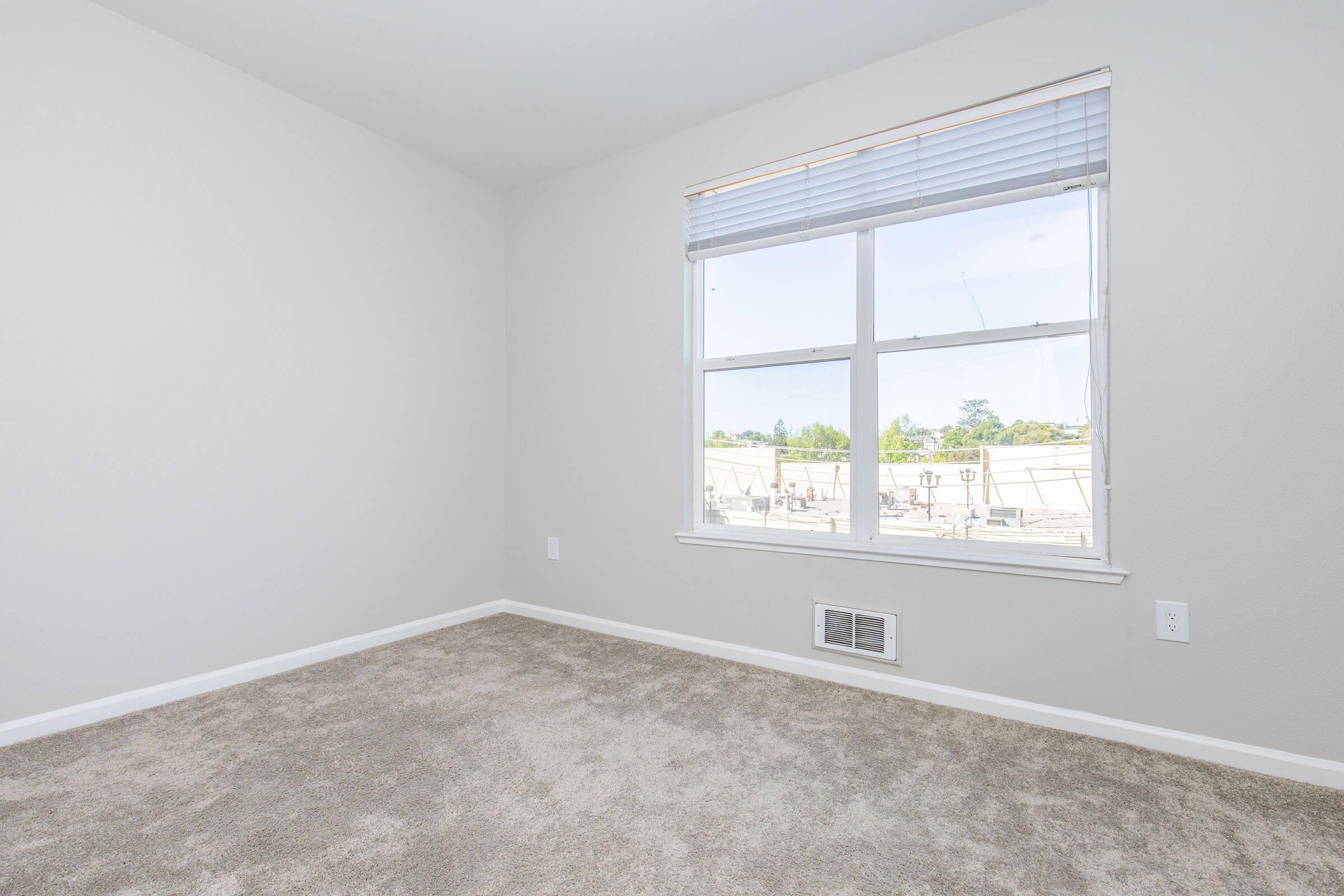 A partially empty room with light gray walls and tan carpet. A large window allows natural light to enter, showing an outdoor view. The corner of the room features a wall vent, and the overall space appears clean and ready for furniture placement.