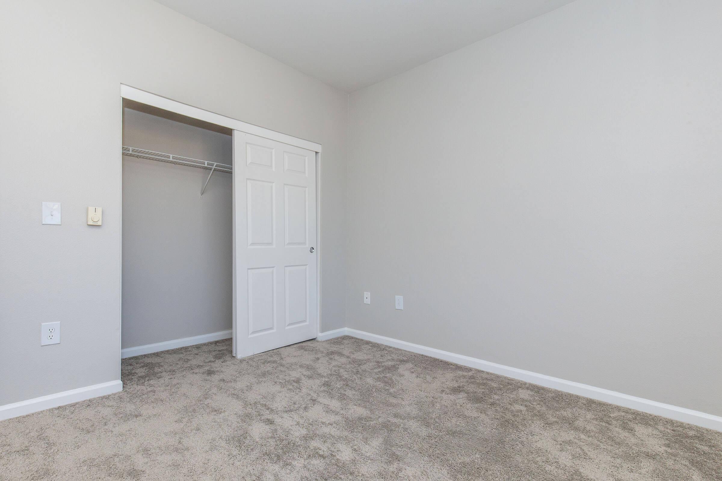 A clean, empty bedroom featuring a light gray wall and beige carpet. There is a white door leading to a closet on the left side, and the room has minimal furnishings, creating an open and airy feel.