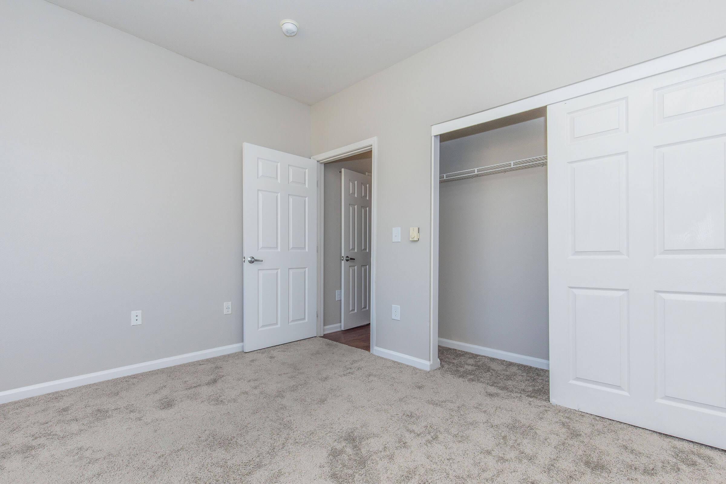 Empty room featuring light-colored walls and carpeted flooring. Two white doors are visible: one leads to a closet with a shelf, and the other opens to a hallway. The room has a bright and clean appearance, ideal for various purposes such as a bedroom or home office.
