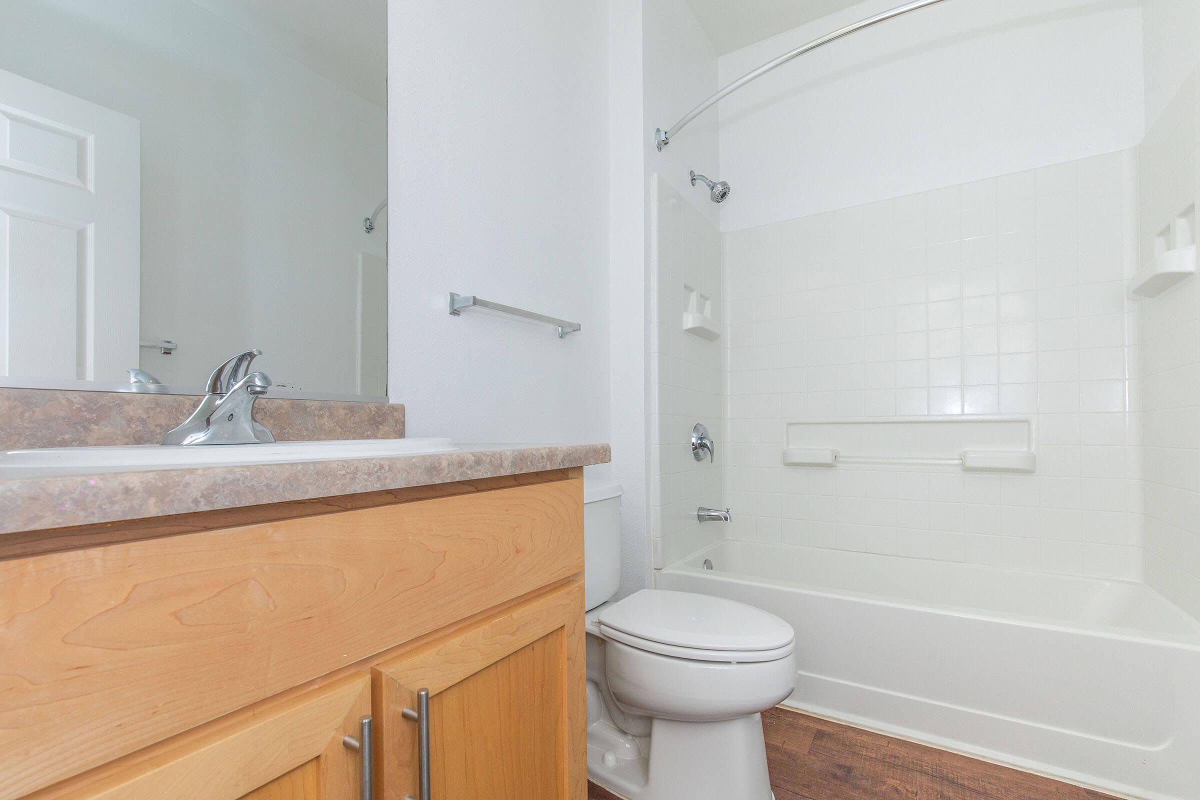 A clean, modern bathroom featuring a bathtub with a showerhead, a sink with a faucet, and wooden cabinetry. The walls are white, and the floor has a wood-like finish. Natural light is coming from an unseen window or door, creating a bright and inviting atmosphere.