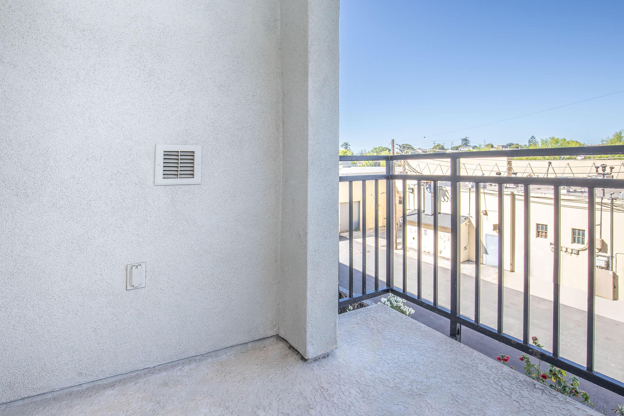 A balcony view with a light-colored wall and a railing. The floor is plain, and there is a small vent or outlet on the wall. Beyond the balcony, a clear blue sky is visible, along with rooftops and structures in the background. Some greenery is also present.