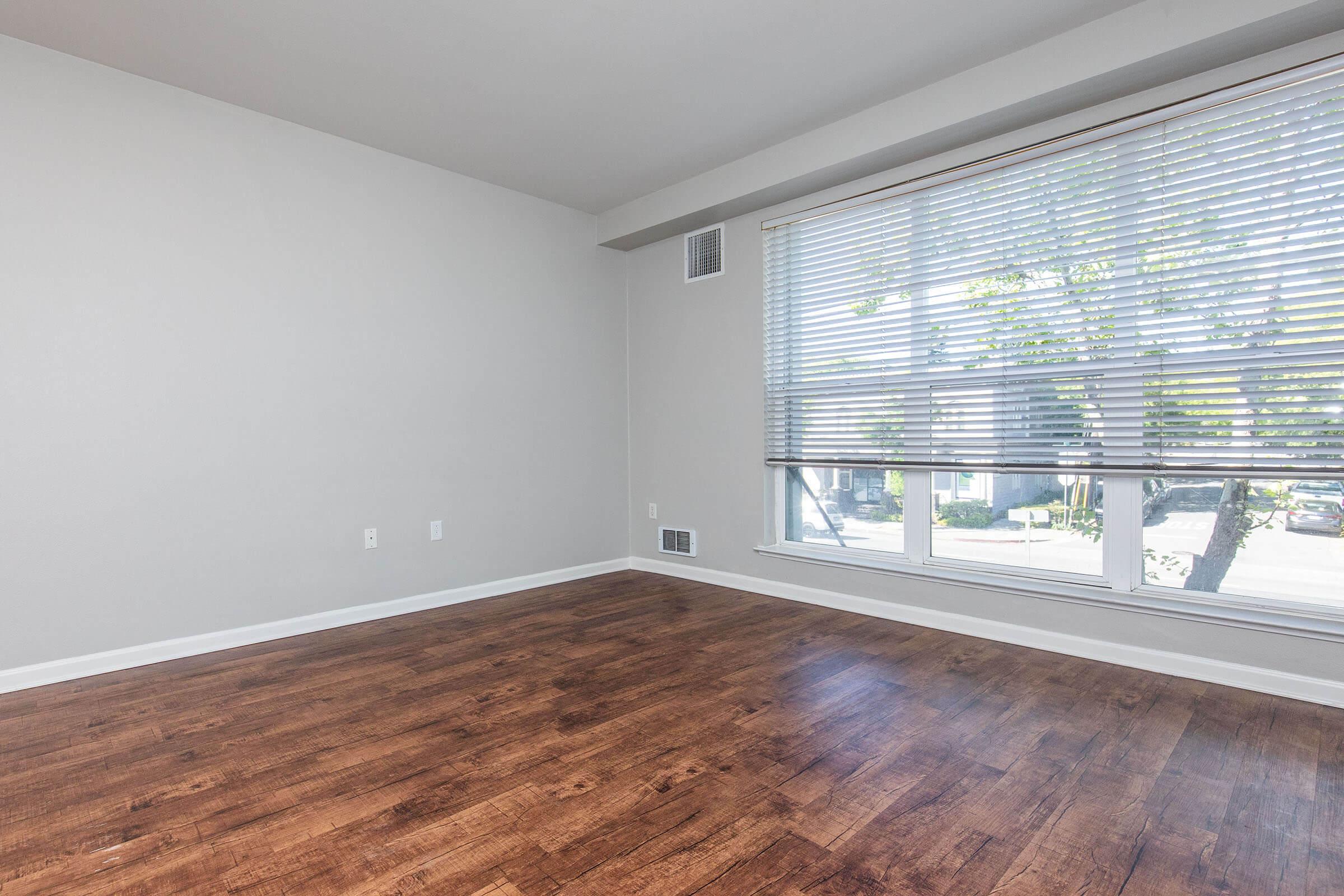 Empty room with light-colored walls and a large window covered by white blinds. The floor is made of dark wood. Natural light is streaming through the window, illuminating the space. There are no furniture or decorations present, creating a minimalist and spacious feel.