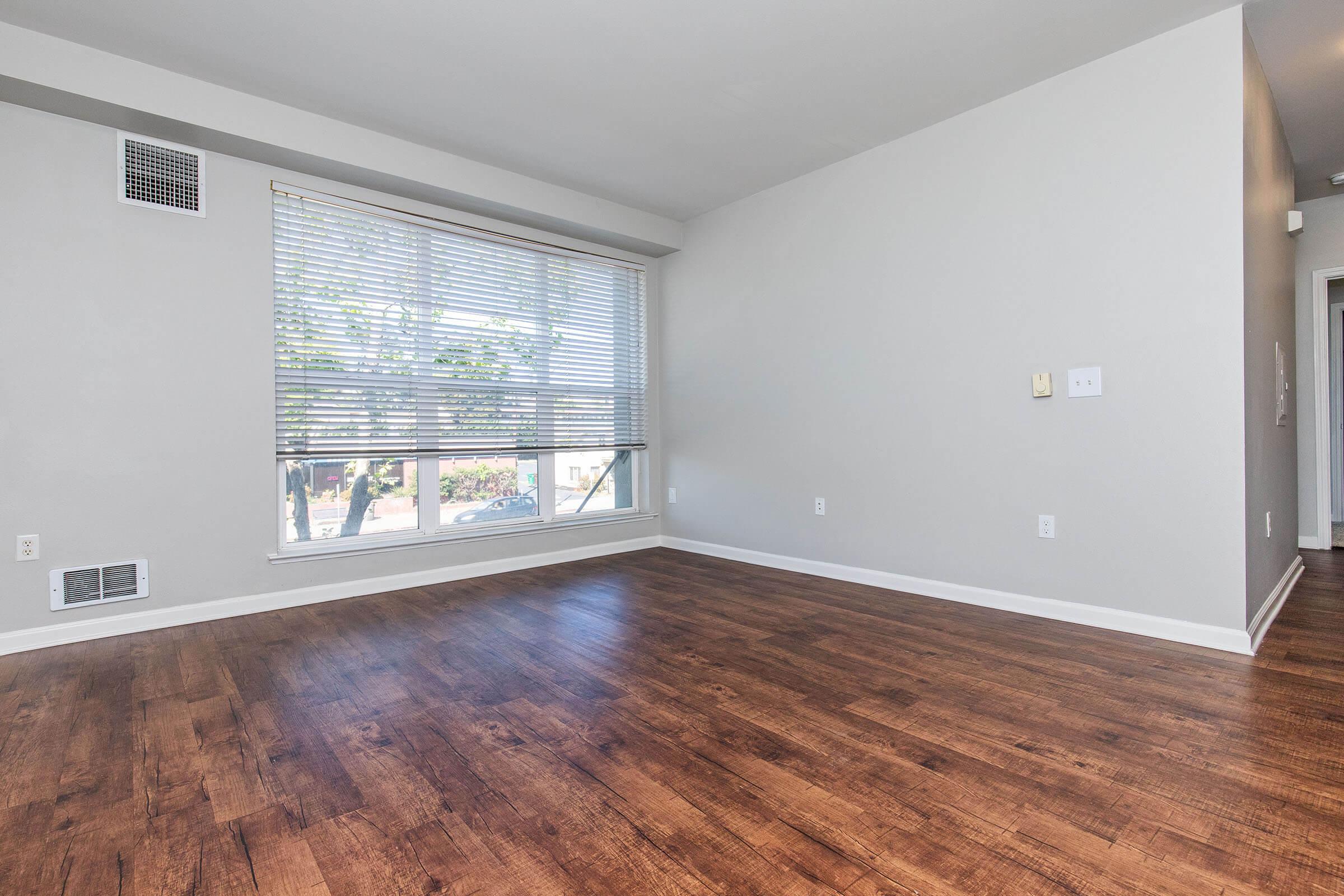 A spacious and well-lit room with light gray walls and hardwood flooring. A large window with white blinds allows natural light to enter, and there is a small wall vent. The room is empty, providing a blank canvas for furnishing.
