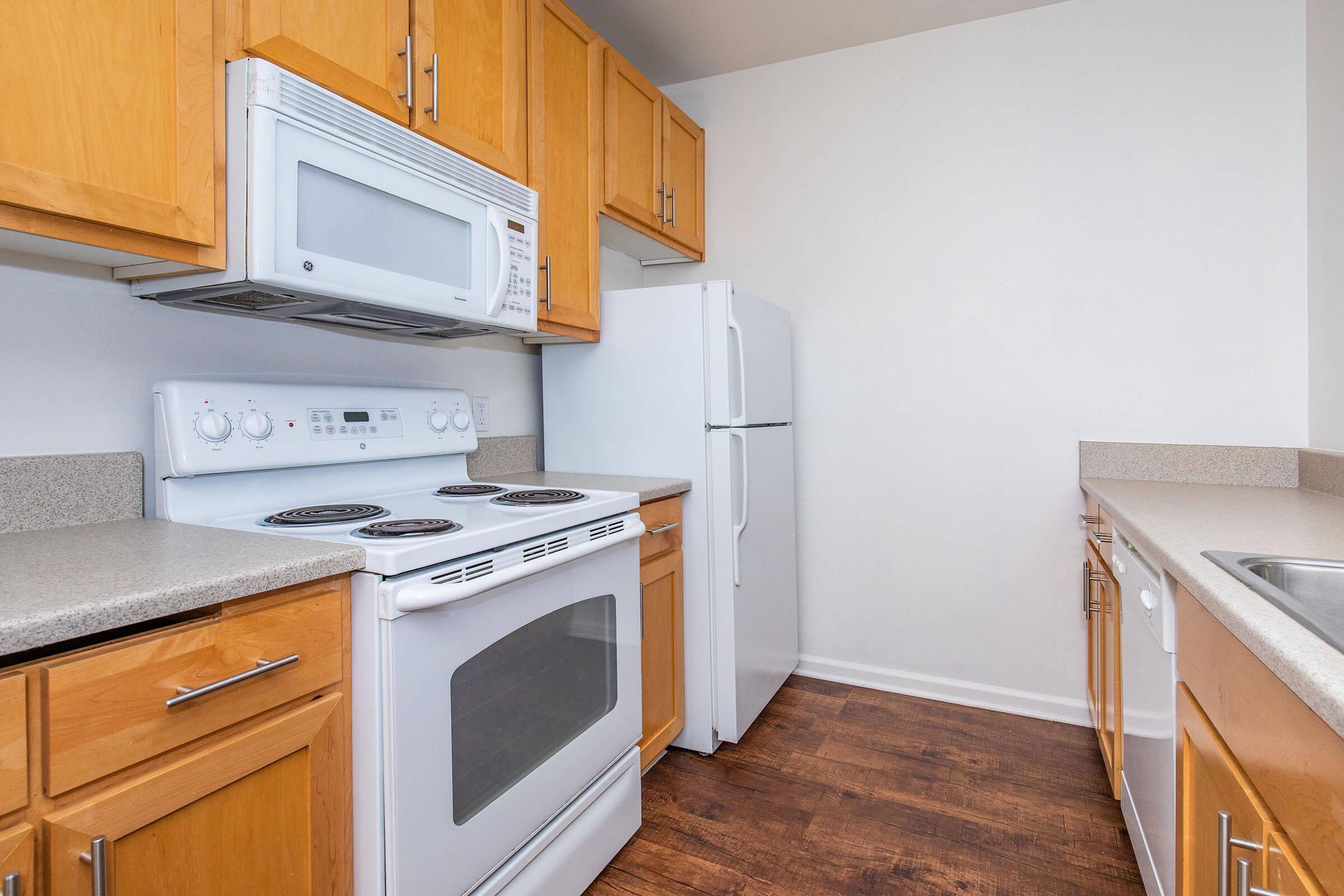 A modern kitchen featuring light wooden cabinets, a white oven with an integrated microwave above, a white refrigerator, and a countertop with a beige surface. The flooring is dark wood, and the space is well-lit with neutral wall colors, creating a clean and inviting atmosphere.