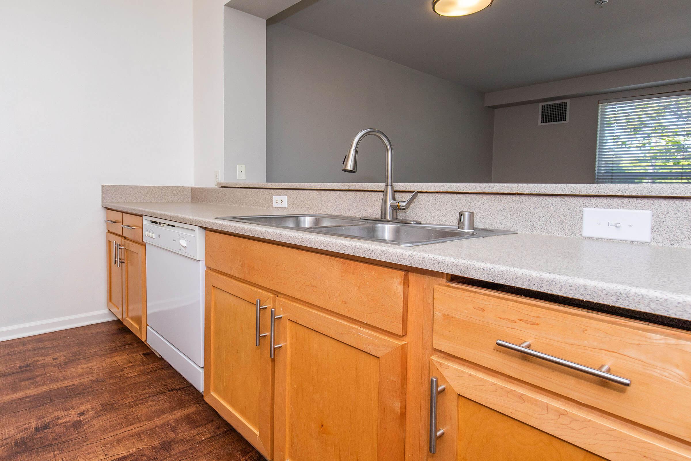 Modern kitchen featuring wooden cabinets, a double sink, a dishwasher, and a light-colored countertop. Natural light comes through a window in the background, illuminating the space. The flooring is a rich, dark wood, adding warmth to the area.
