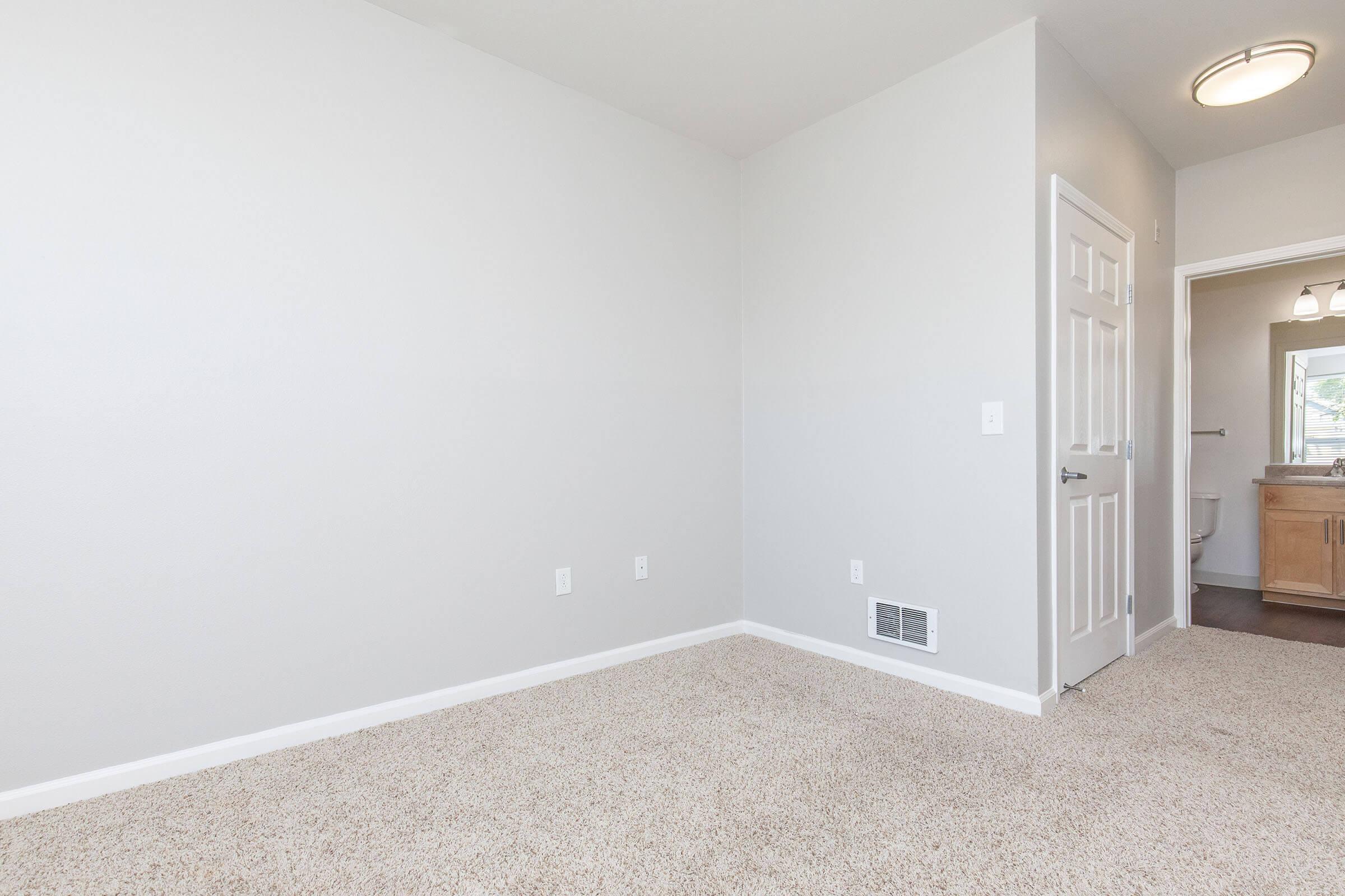 A vacant room with light gray walls and beige carpet. There is a door on the left leading to another room, and a doorway at the far end that appears to lead to a bathroom. A ceiling light is visible overhead, providing illumination in the space.