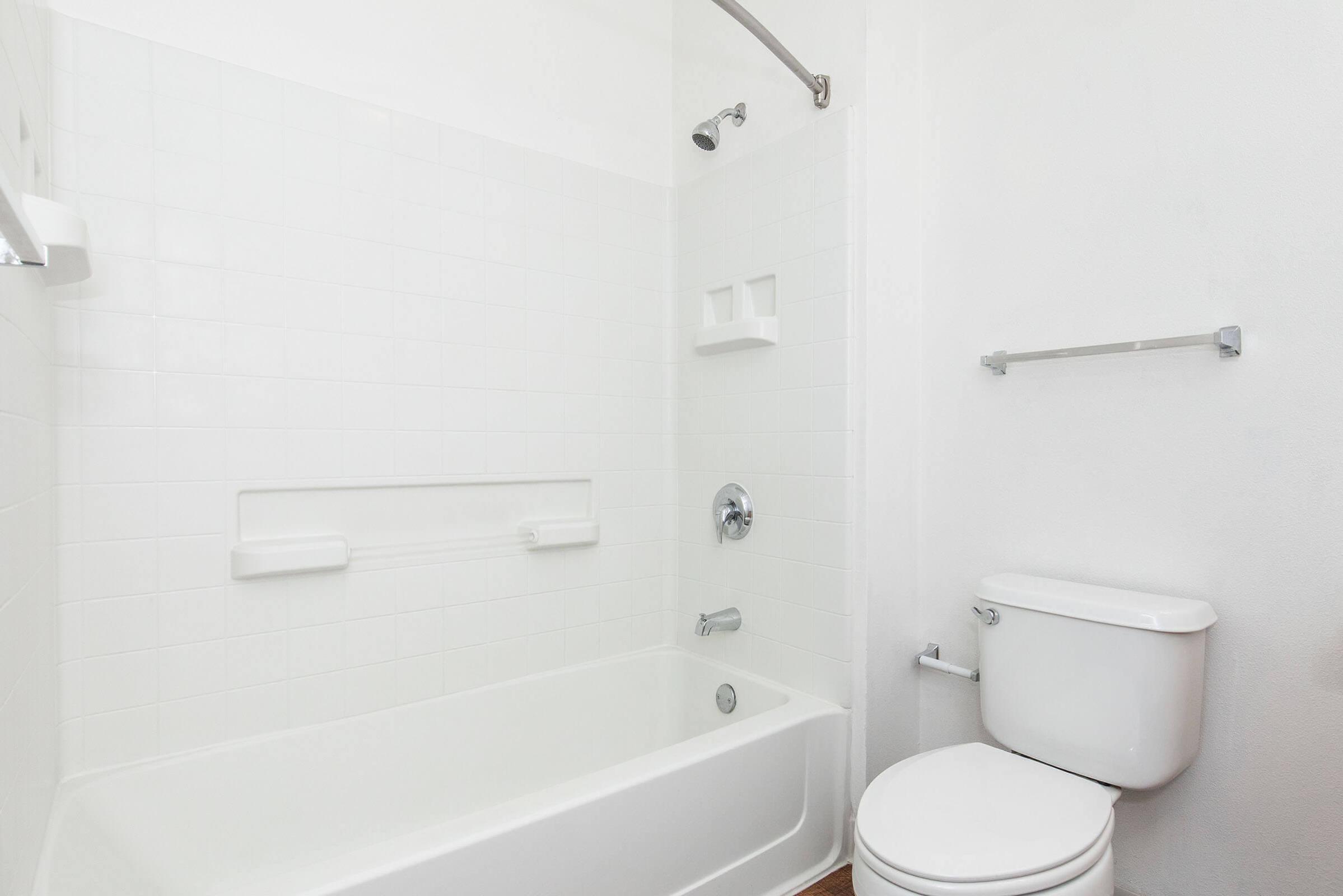 A clean and modern bathroom featuring a white bathtub with a shower, white tiled walls, a toilet, and minimalistic fixtures. The space is well-lit with a bright and airy feel, showcasing a simple and functional design.