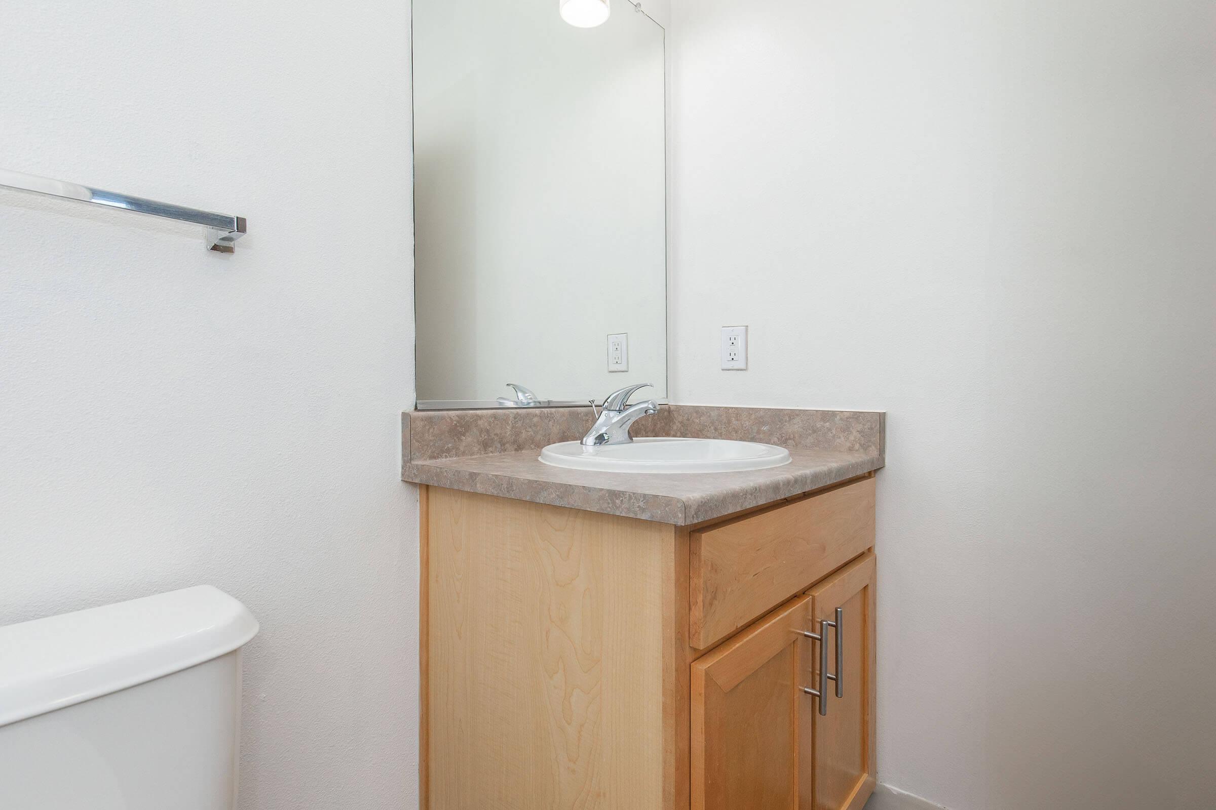 A clean and simple bathroom setting featuring a light-colored wall, a wooden vanity with a sink and countertop, a mirror above the sink, and a white toilet. The overall look is modern and minimalistic, with a focus on functionality.