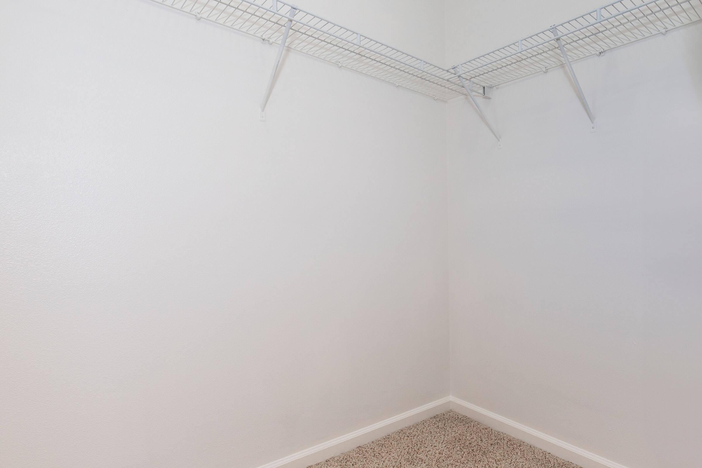 A small, empty closet featuring plain white walls and a carpeted floor. Two wire shelves are mounted on the walls, providing storage space, but the closet is otherwise unoccupied and minimalistic in design.