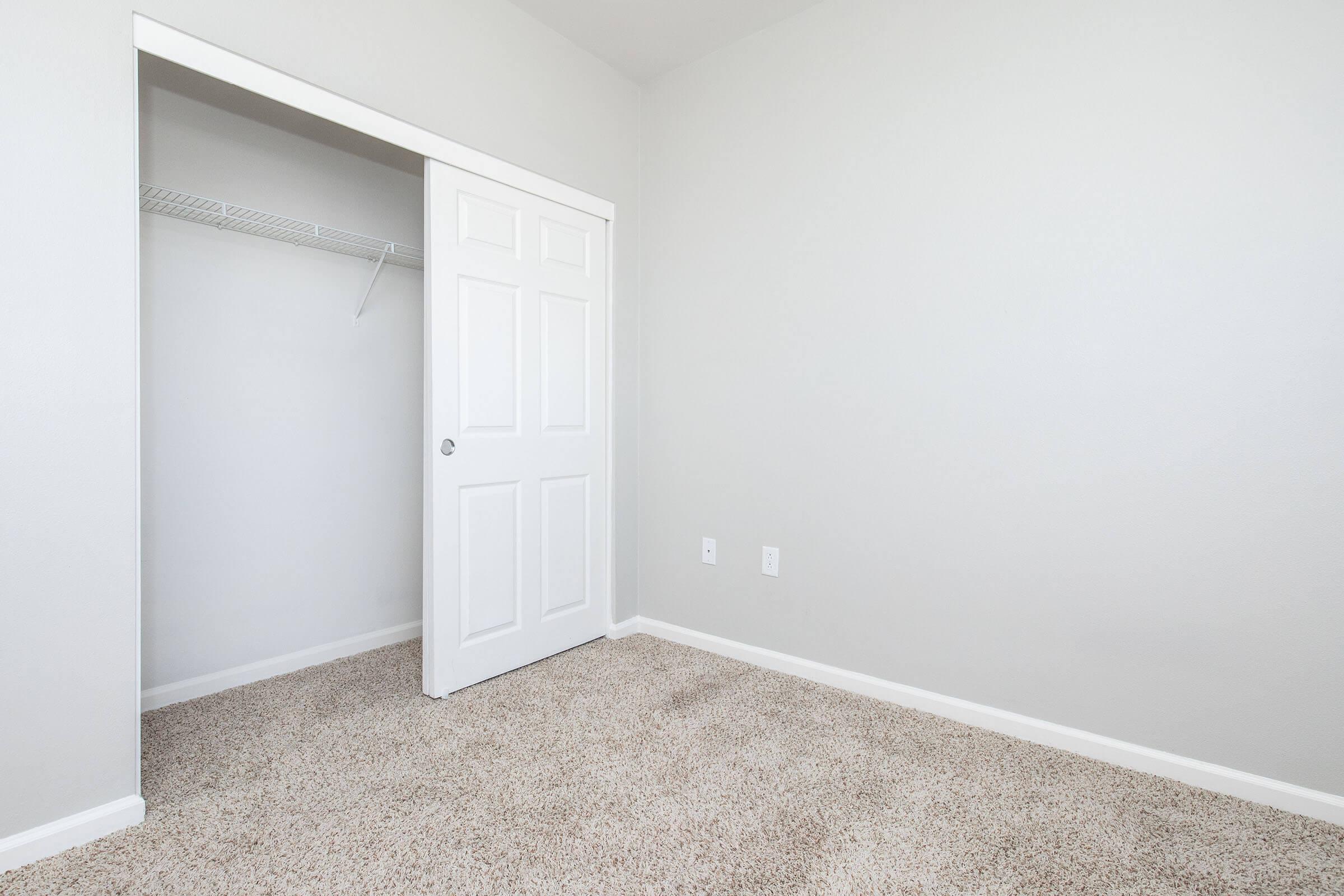 A small, empty room featuring a light gray wall and beige carpet. A white door is partially open, leading to a closet with a shelf. The room has no furniture, creating a minimalistic and spacious feel.