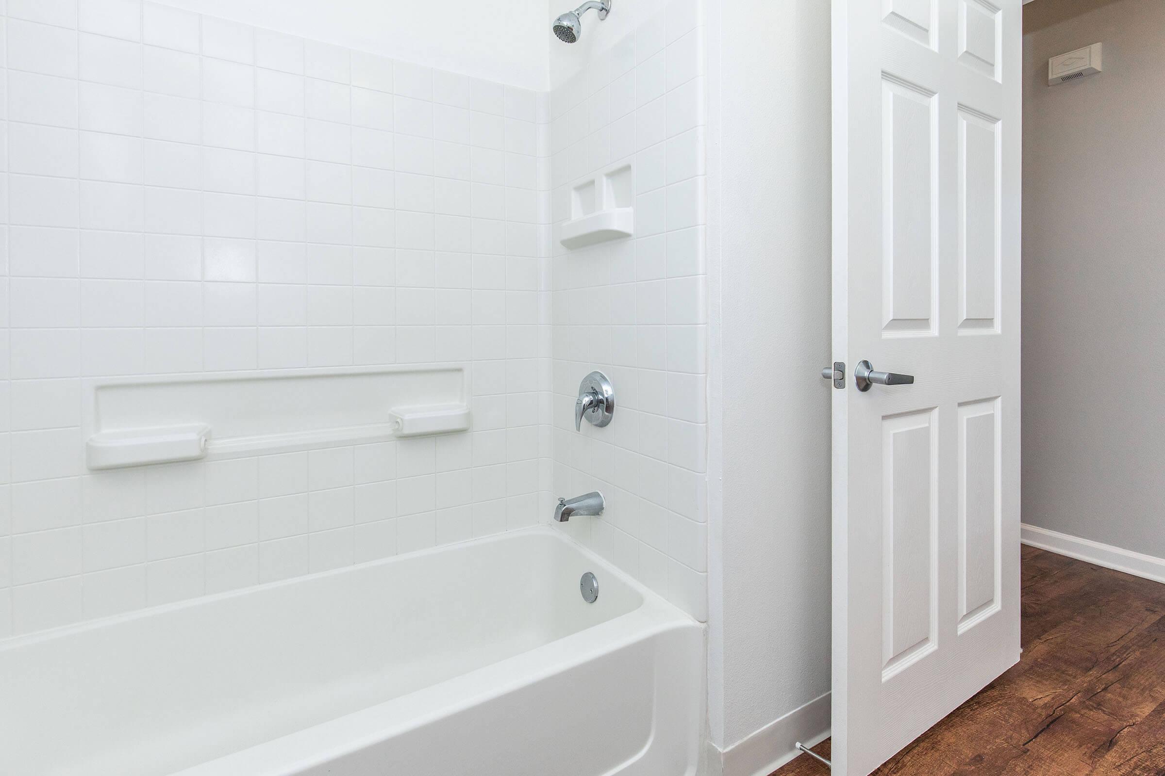 A clean, modern bathroom featuring a white bathtub and shower with white tiles. There are two built-in shelves on the wall for storage, and a door leading to another room. The flooring is a warm wood tone, adding contrast to the bright white walls and fixtures.