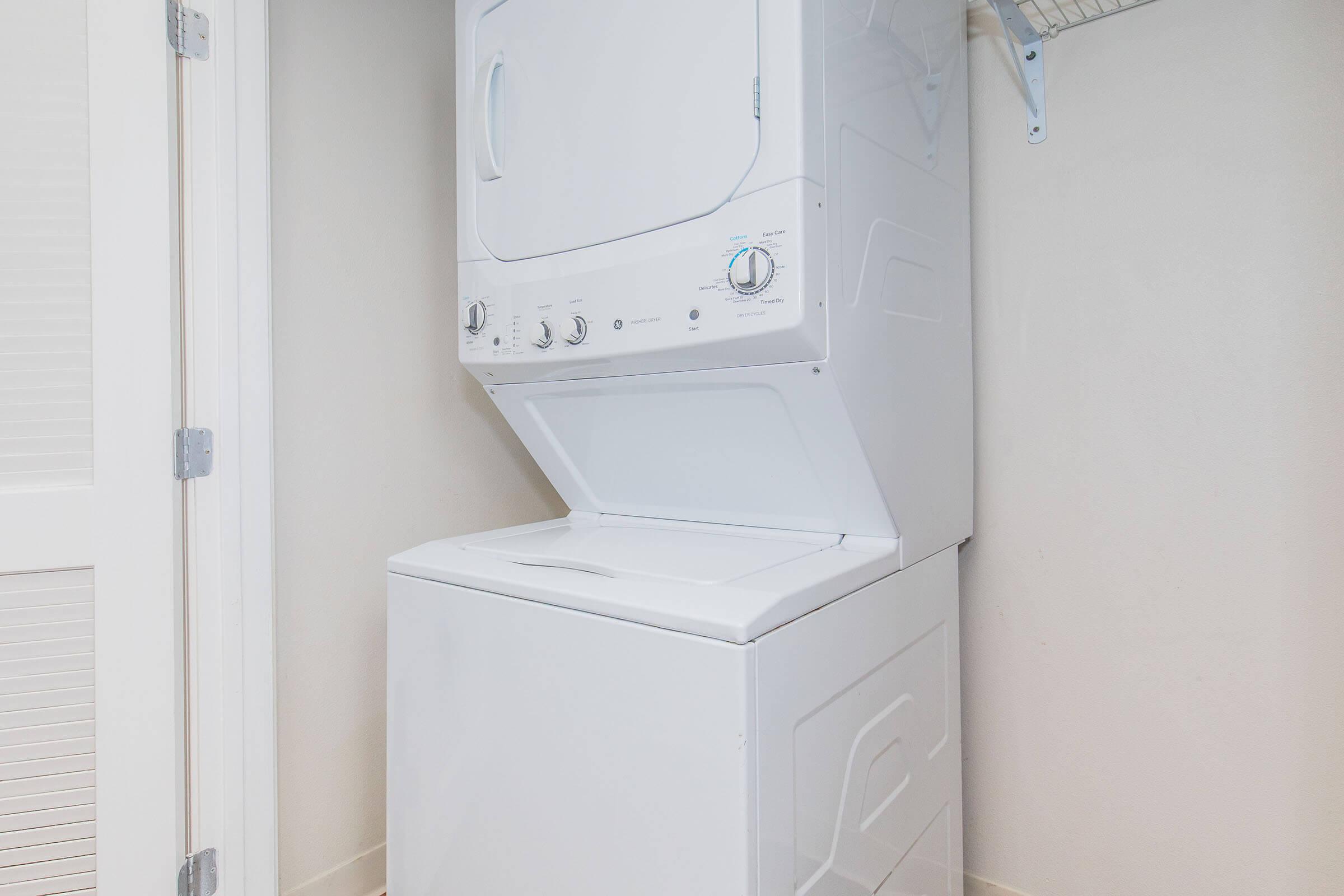 A stacked washer and dryer set in a light-colored laundry room with a smooth wall and a shelf above for storage. The appliances are white and compact, suited for small spaces.