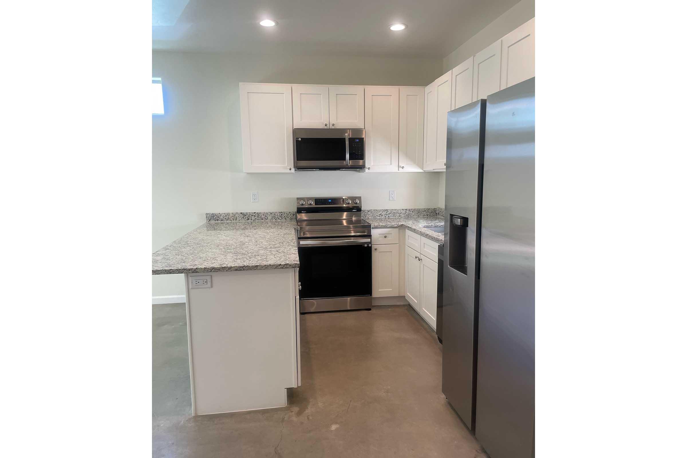 A modern kitchen featuring white cabinetry, a stainless steel refrigerator, an oven with a stovetop, and a microwave. The countertops are made of speckled granite and the floor is a concrete finish. Bright lighting enhances the open, airy feel of the space.