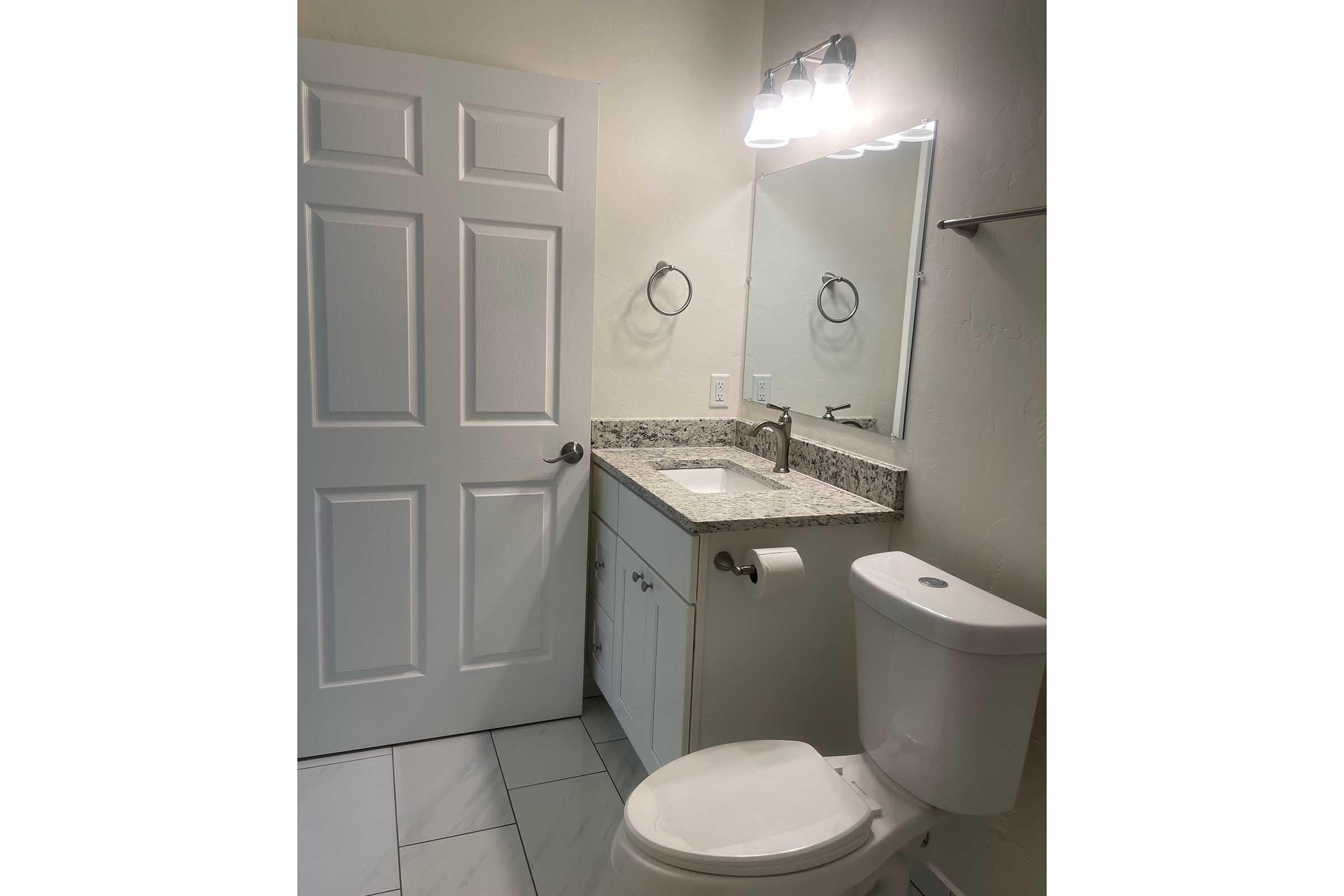 A clean, modern bathroom featuring a white vanity with a granite countertop, a sink, a mirror, and a light fixture above. A toilet is positioned to the right, and a towel ring is mounted on the wall. The floor is tiled, and there is a door leading out of the bathroom.