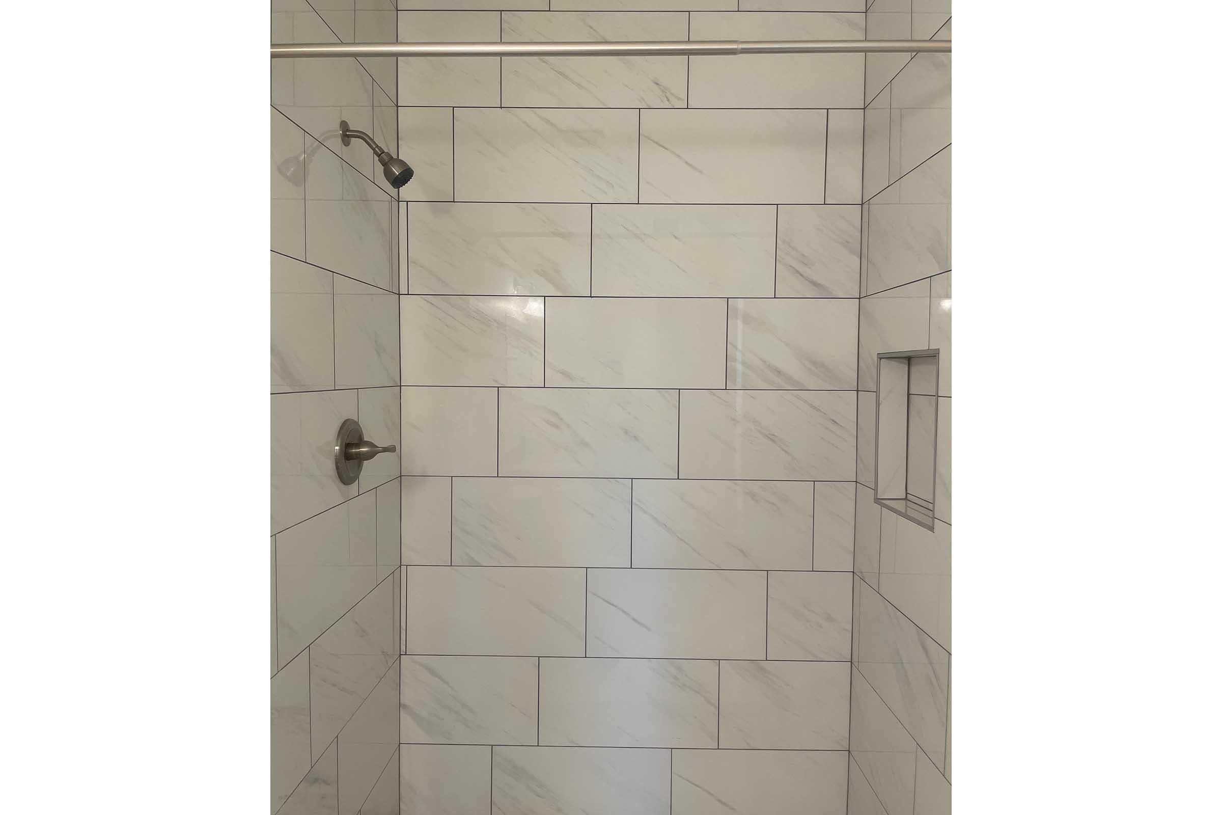 A modern shower space featuring large, light-colored marble tiles on the walls. It includes a chrome showerhead mounted on the wall and a small recess in the tile for storage. The design is sleek and minimalistic, creating a clean and spacious appearance.