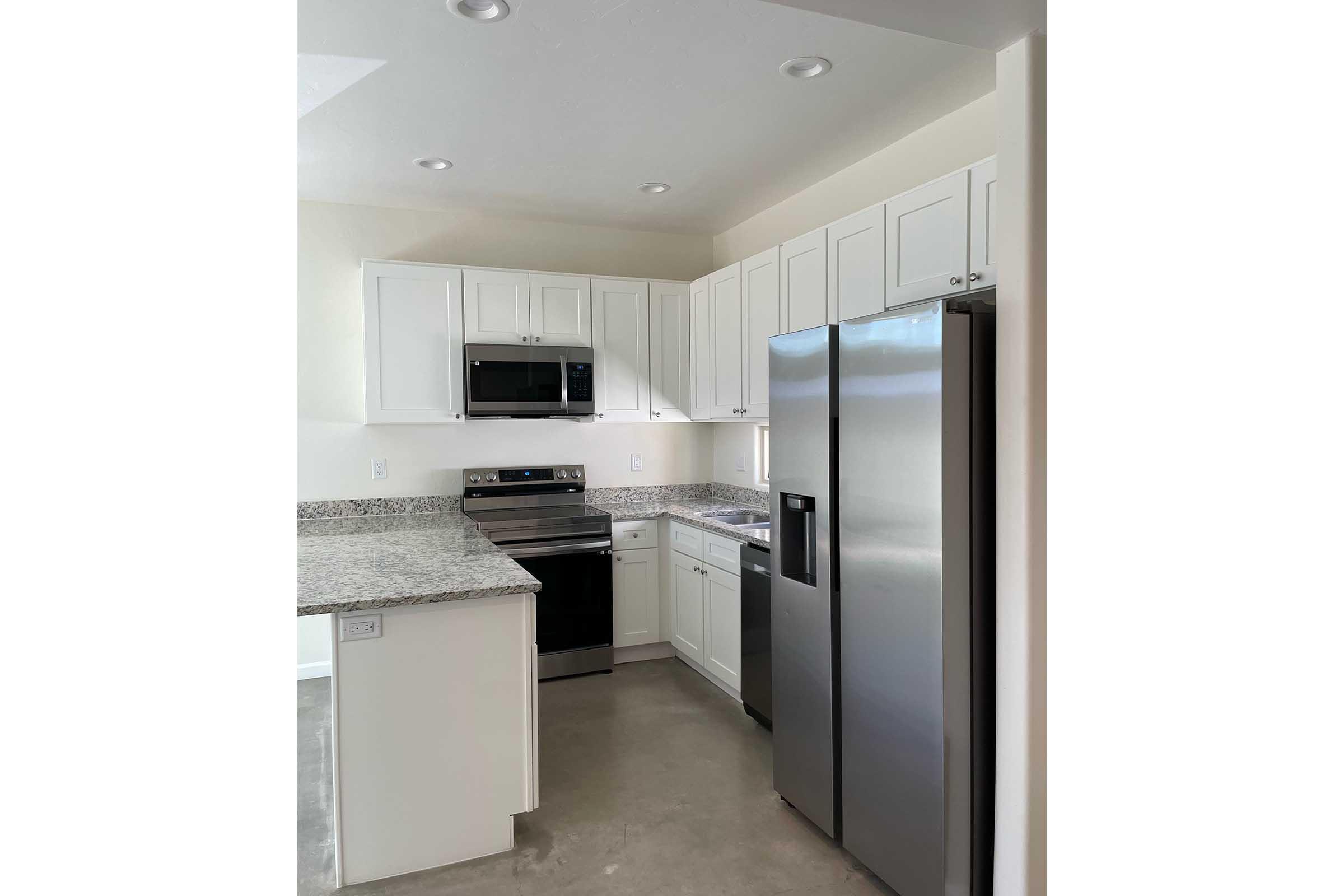 A modern kitchen featuring white cabinetry, granite countertops, stainless steel appliances including a microwave, oven, and refrigerator, with an open layout and natural light from a window. The floor is a light-colored concrete.