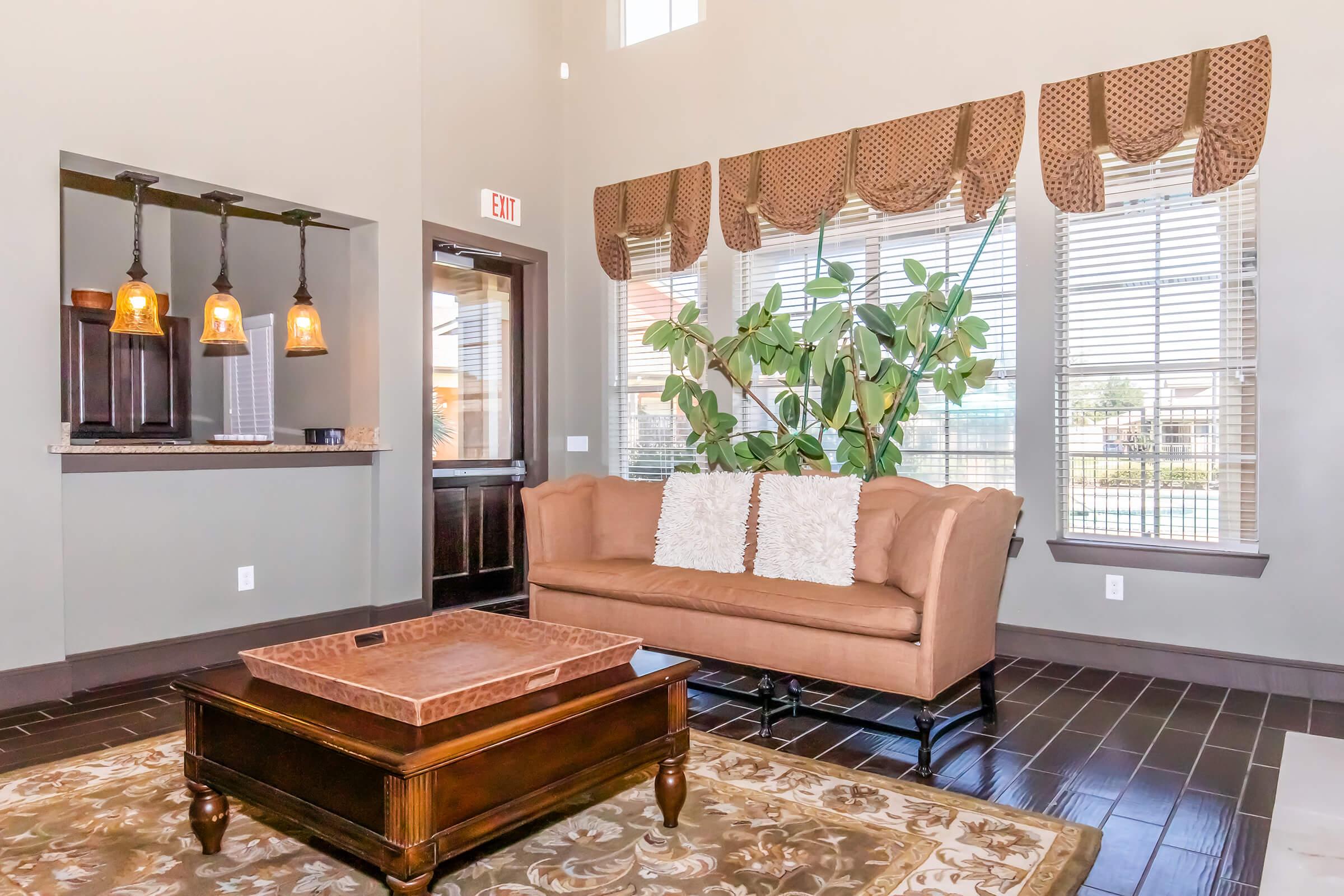 A modern living space featuring a light brown sofa with two cushions, a wooden coffee table on a decorative rug, and large windows with elegant valances. A potted plant adds greenery, and there's a bar area in the background with hanging pendant lights. The walls are painted in neutral tones, creating a warm ambiance.