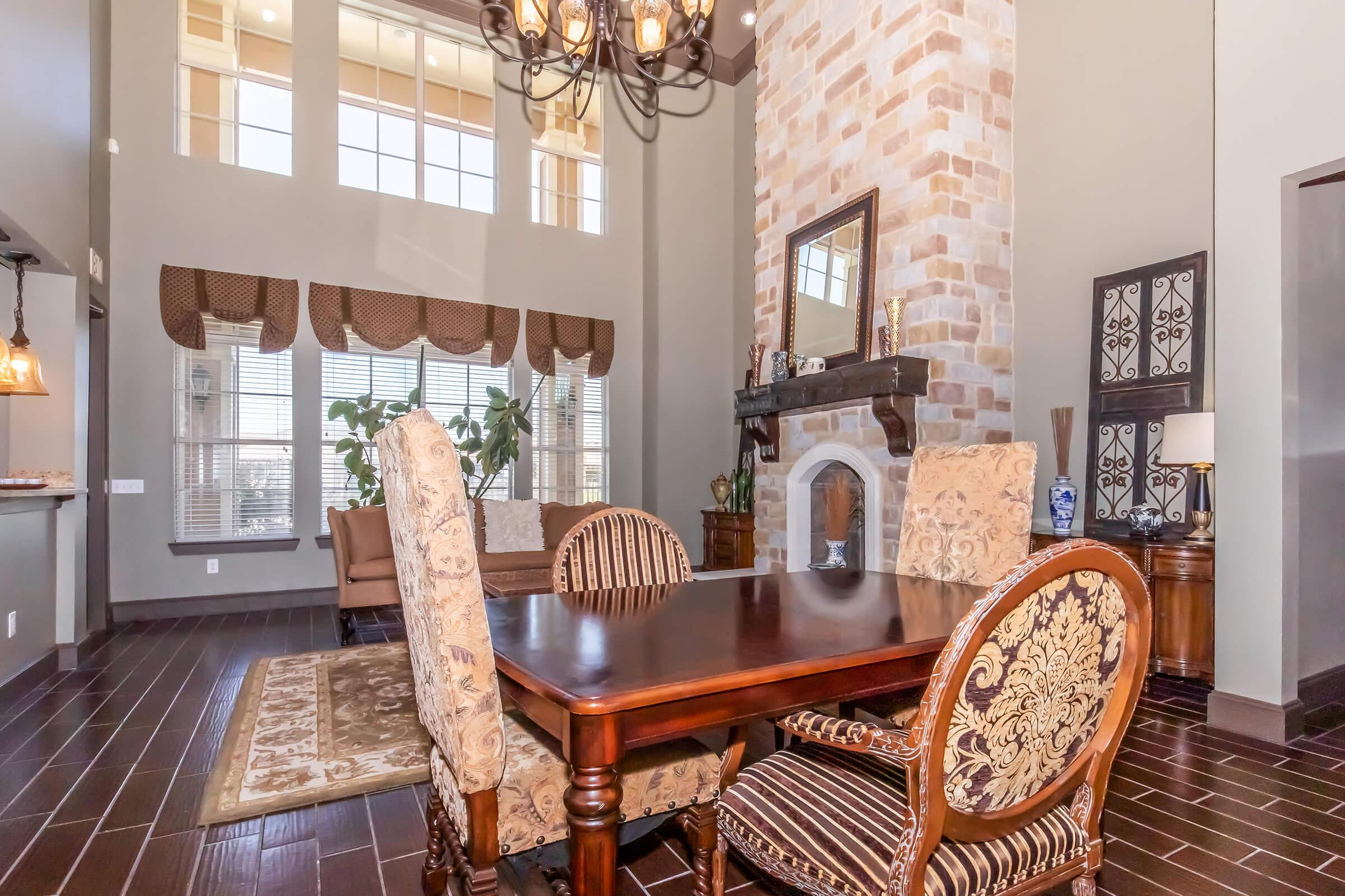 A cozy dining area featuring a dark wooden table surrounded by ornate chairs, with a stylish chandelier overhead. The room has large windows allowing natural light, stone accents on the fireplace, and decorative elements like plants and artworks, creating an inviting atmosphere.