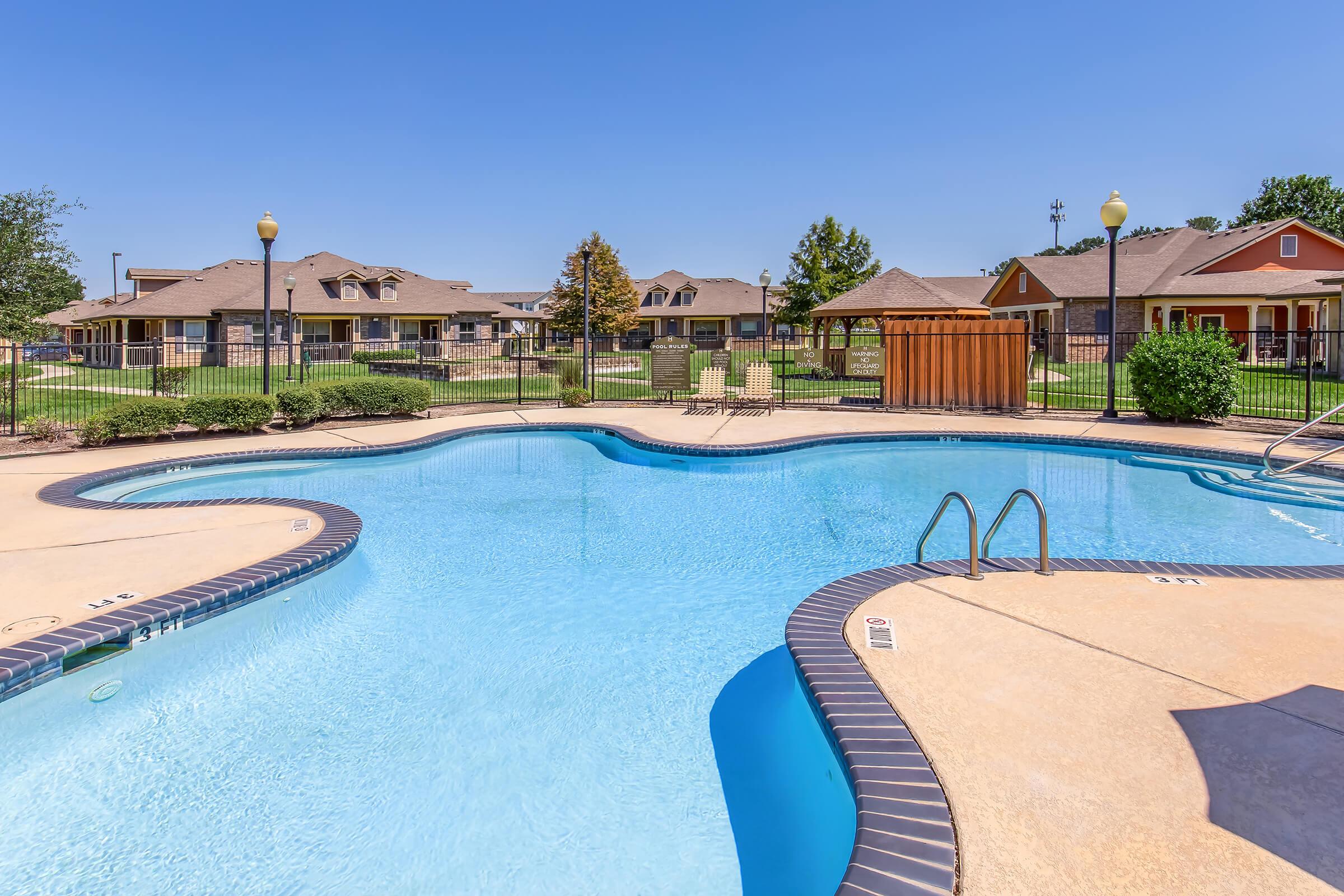 A clear blue swimming pool surrounded by a well-maintained landscape, including green grass and trees. In the background, there are residential buildings with a sunny sky above. The pool features a curved design and steps leading into the water, with a safety fence and lounge areas visible nearby.