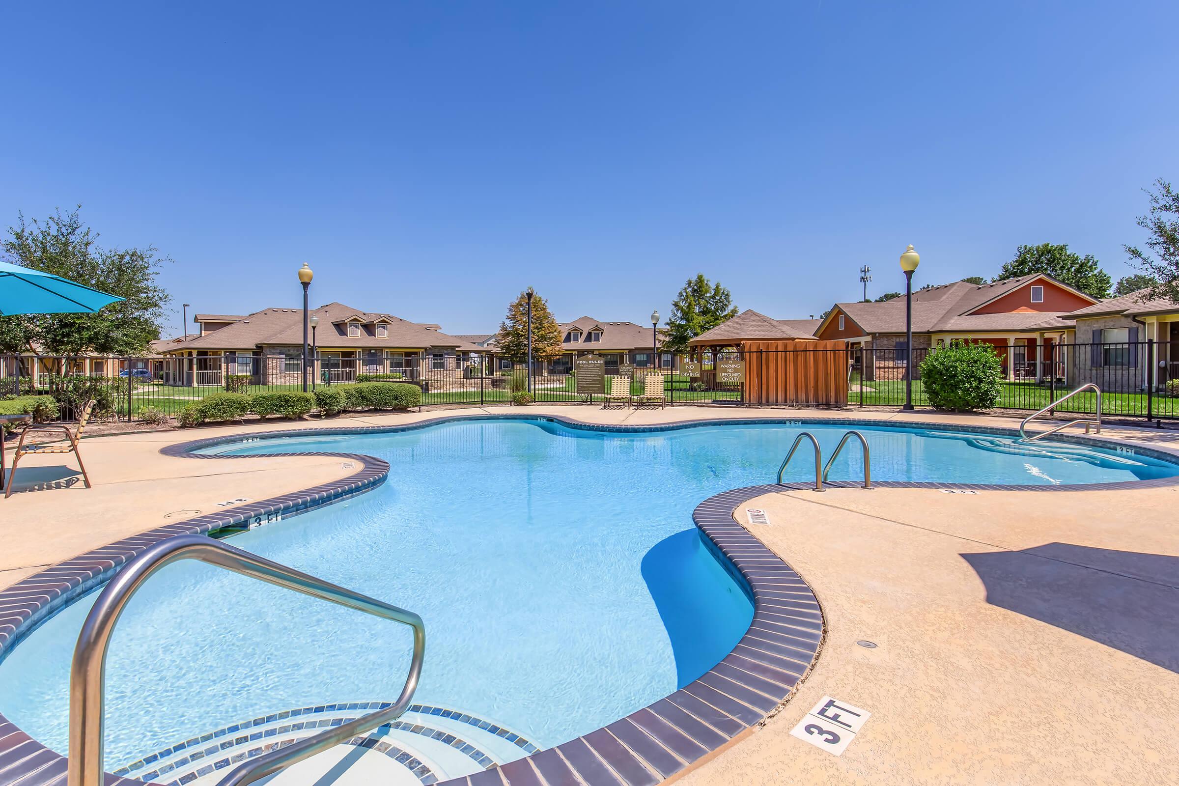 A clean, blue swimming pool with a gradual slope, surrounded by lounge chairs and umbrellas. In the background, residential buildings are visible with well-maintained lawns and trees under a clear blue sky. The area appears inviting and ready for relaxation or leisure activities.