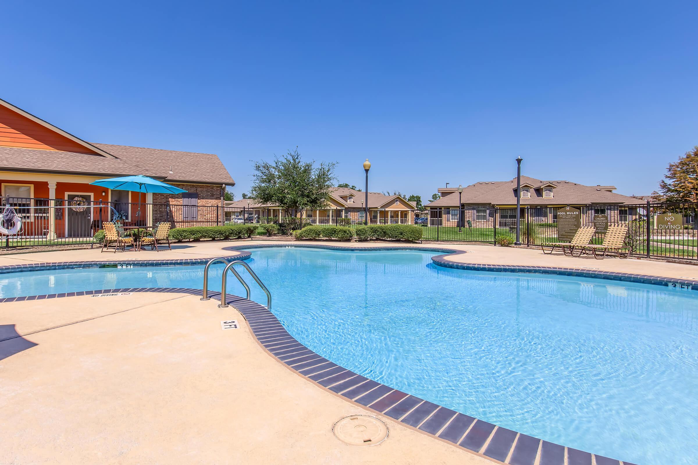 A sparkling blue swimming pool surrounded by a spacious deck, with lounge chairs and umbrellas. In the background, there are well-maintained residential buildings and green trees under a clear blue sky. The scene conveys a relaxing outdoor atmosphere perfect for leisure.