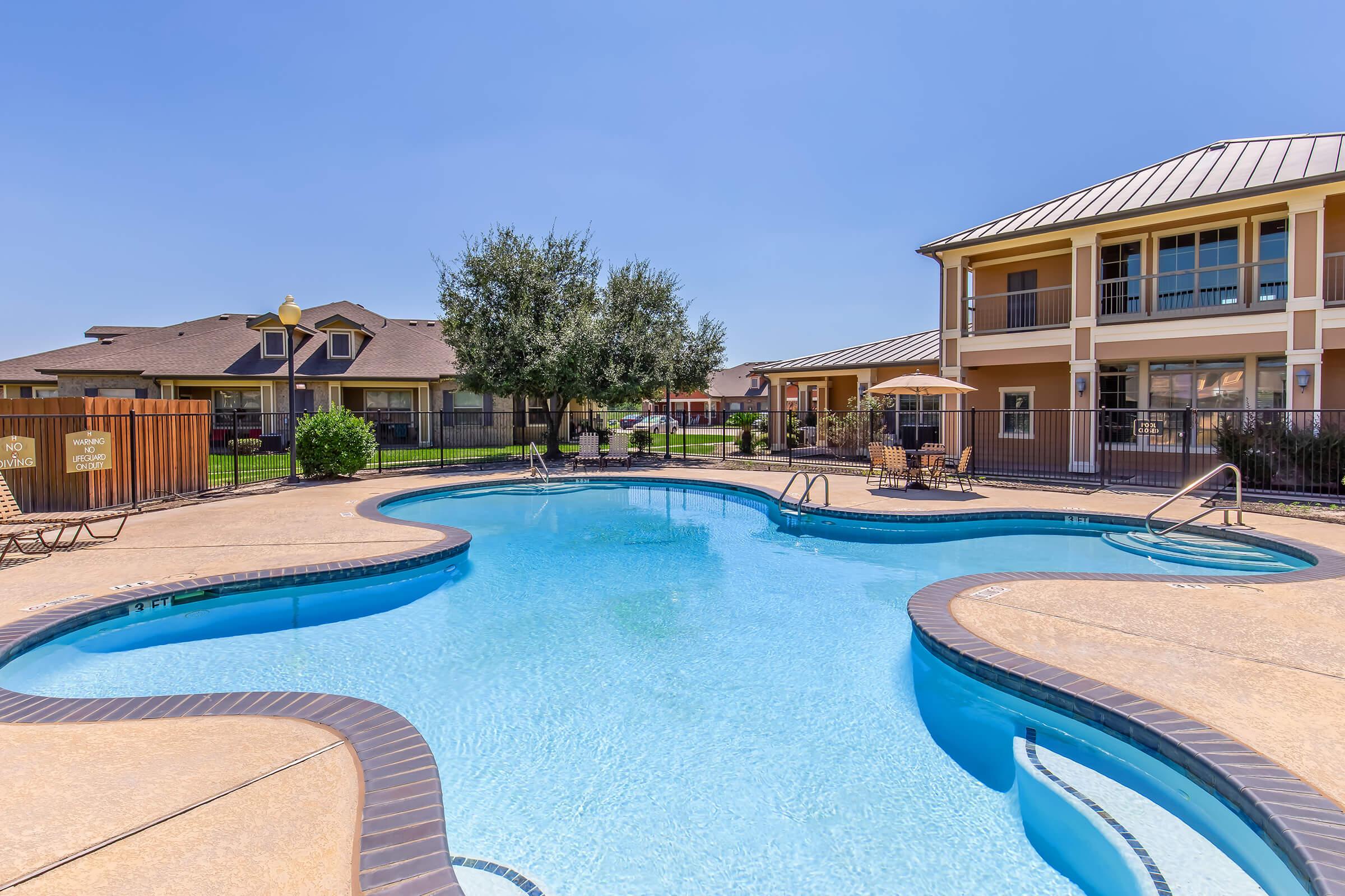 A clear blue swimming pool with curved edges, surrounded by lounge chairs and a grassy area. In the background, there are residential buildings with balconies and trees, under a bright blue sky.