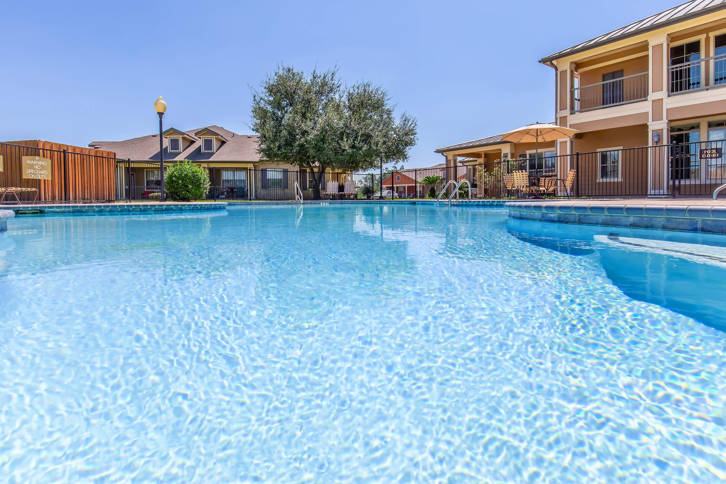 A clear blue swimming pool surrounded by a patio area, with lounge chairs and umbrellas. In the background, there are two residential buildings with a well-maintained lawn and trees providing shade. The sky is blue and clear, creating a relaxing atmosphere.