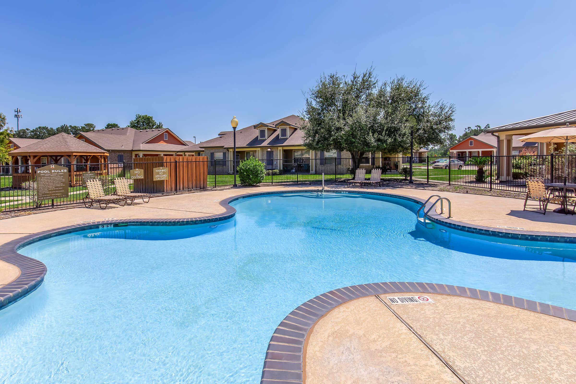 A sparkling blue swimming pool shaped like a kidney, surrounded by lounge chairs. In the background, there are residential buildings and a gazebo area. The scene is sunny with a clear blue sky, creating a relaxing atmosphere for guests.