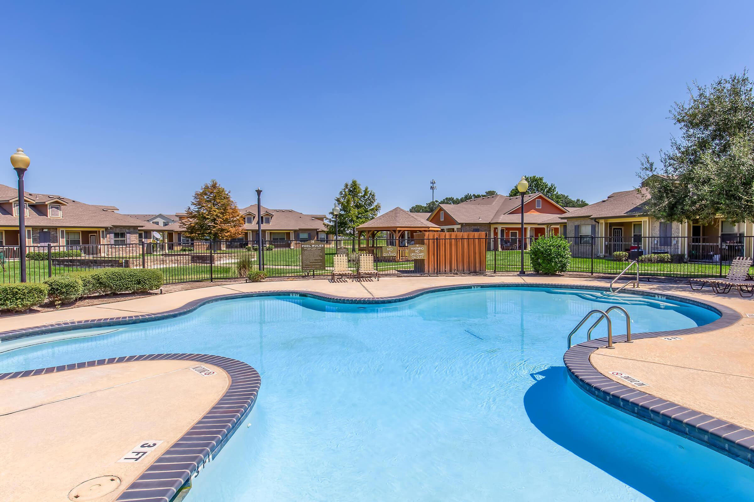 Swimming pool with a clear blue water and lounge chairs surrounding it. The area is well-maintained, featuring greenery and wooden gazebo in the background. Residential buildings are visible in the distance, with a bright blue sky overhead. Ideal for relaxation and leisure.