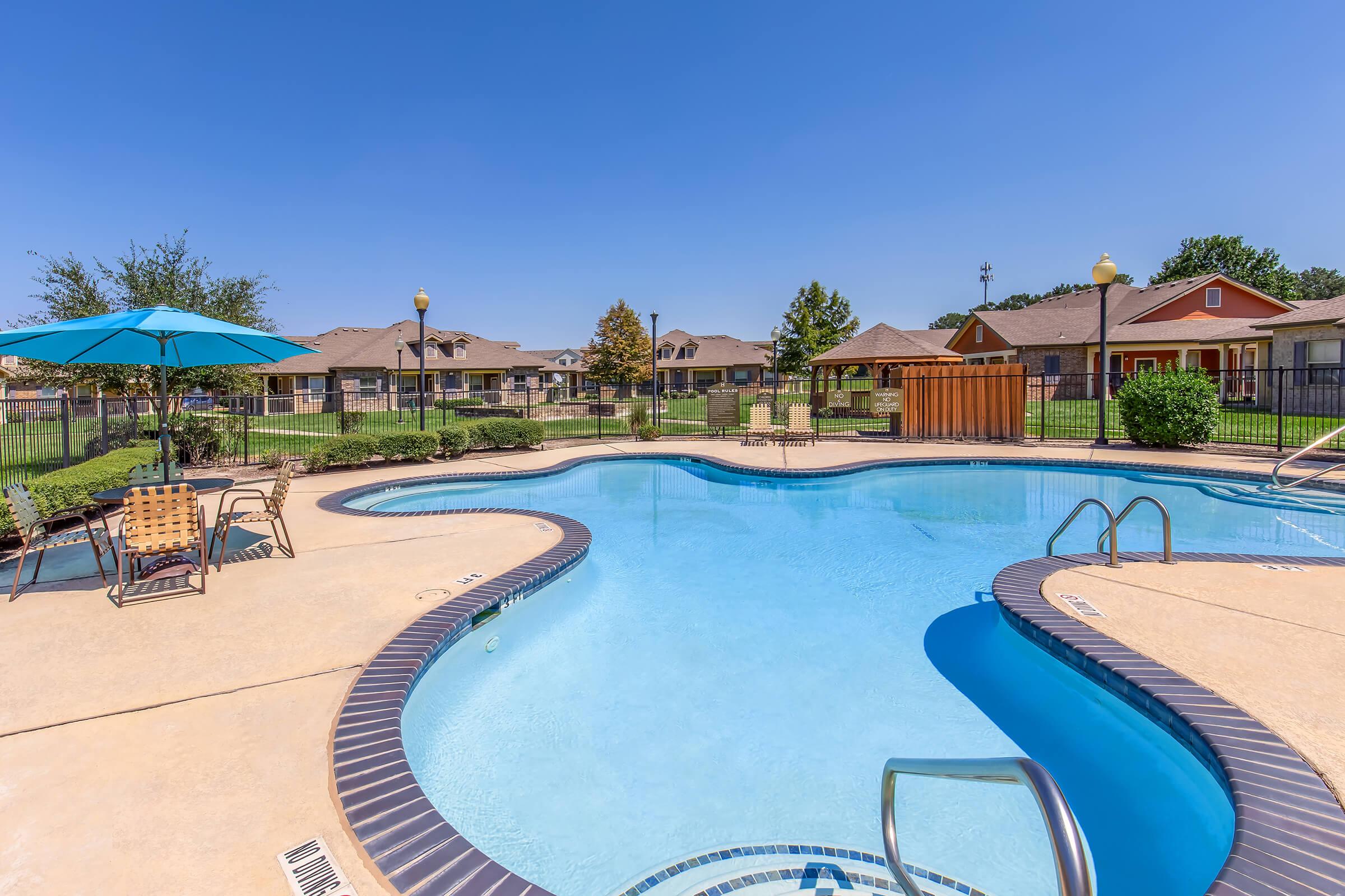A clear blue swimming pool surrounded by a deck with lounge chairs, an umbrella, and landscaped greenery. In the background, there are residential buildings and a fence, under a bright blue sky. The setting appears inviting and well-maintained, ideal for relaxation and enjoyment.