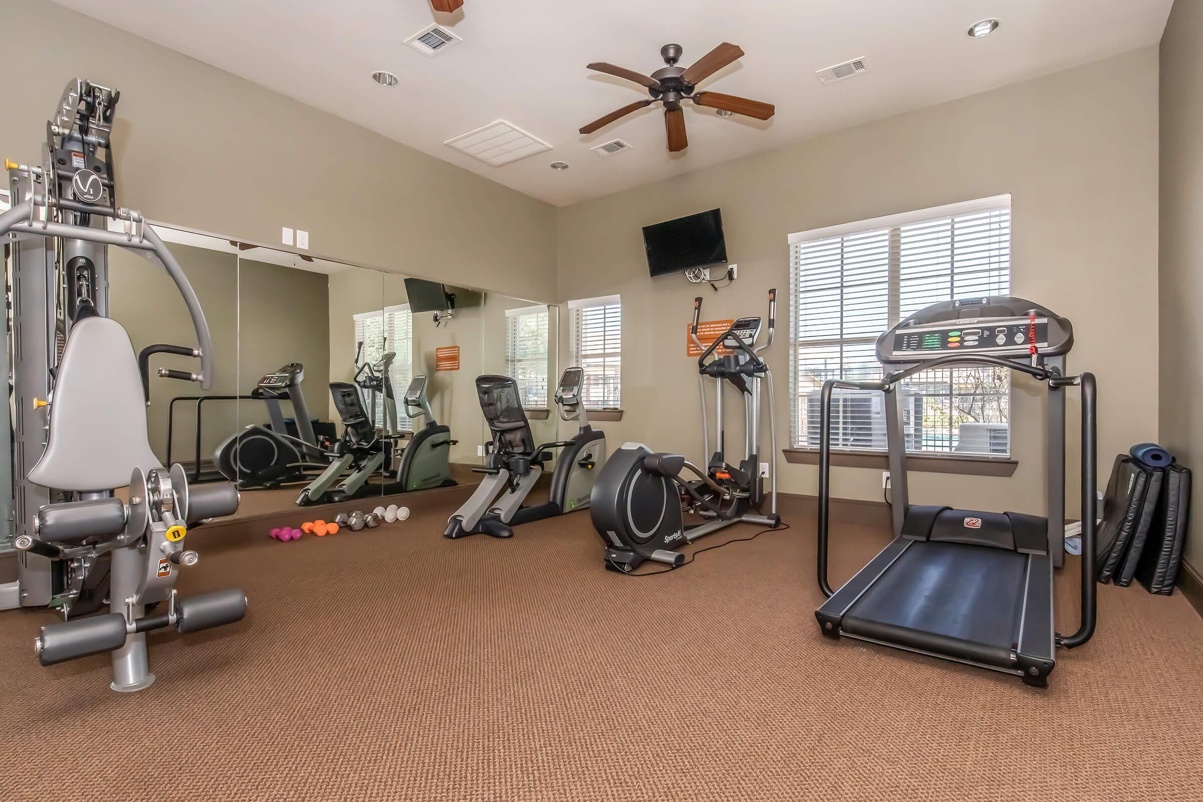 A modern gym interior featuring fitness equipment, including a treadmill, stationary bike, strength-training machine, and free weights. Large mirrors reflect the workout area, which is well-lit with natural light coming from windows. The floor is carpeted, and a ceiling fan adds ventilation.