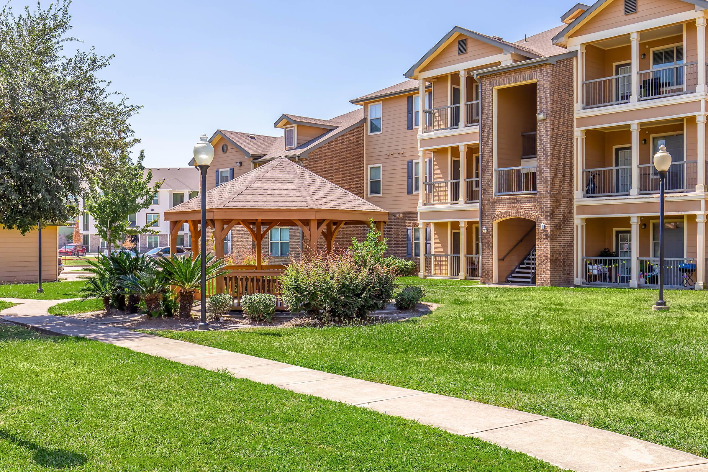 A well-maintained apartment complex featuring a gazebo surrounded by lush green grass and palm trees. The background shows multiple three-story residential buildings with balconies. Clear blue skies provide a bright and sunny atmosphere. A paved walkway leads through the lawn, enhancing the outdoor appeal.
