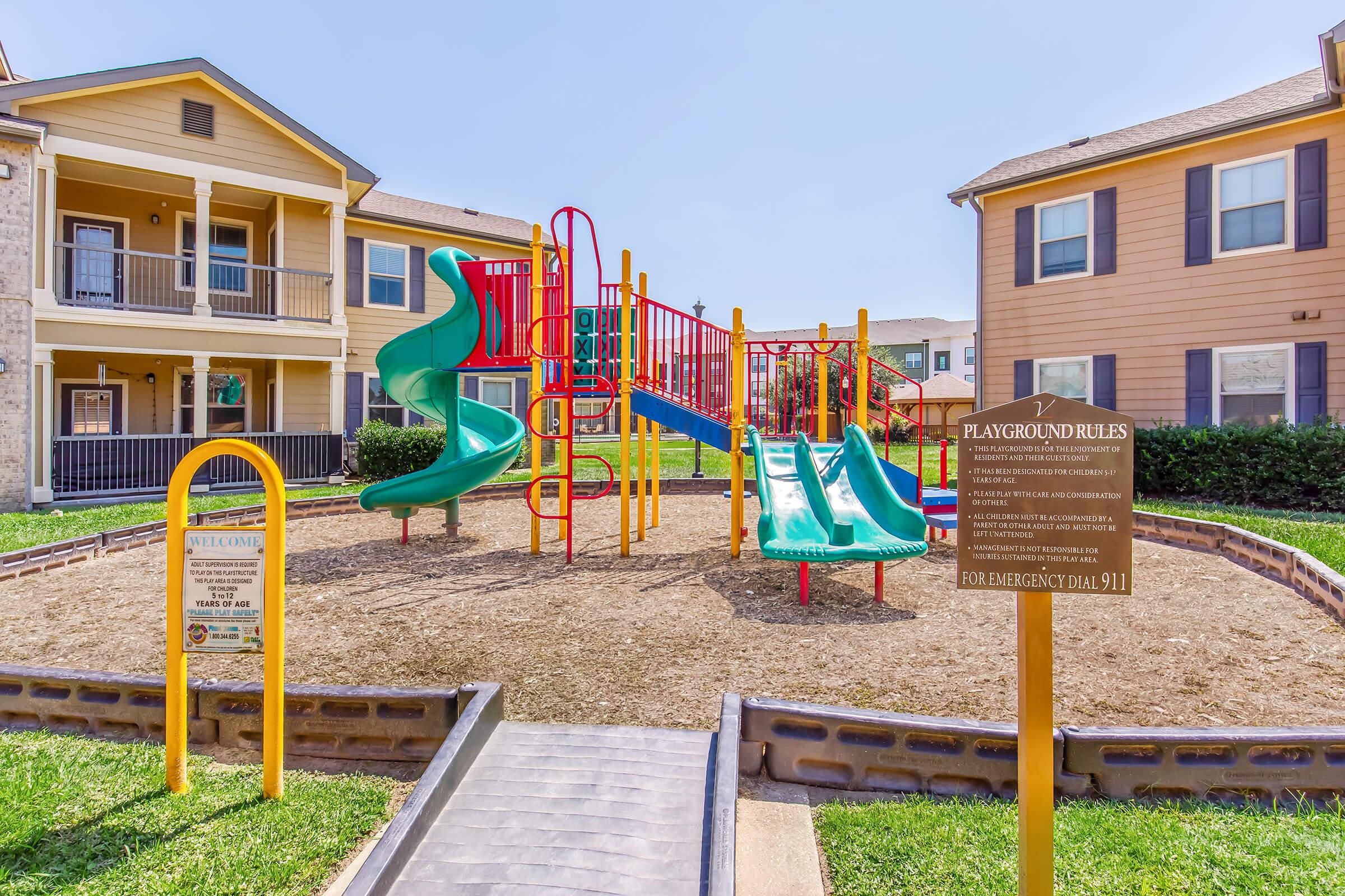 Playground area featuring colorful play structures with slides, surrounded by grassy areas and two apartment buildings in the background. A sign detailing playground rules is visible, along with a pathway leading to the play area. Clear blue sky overhead.