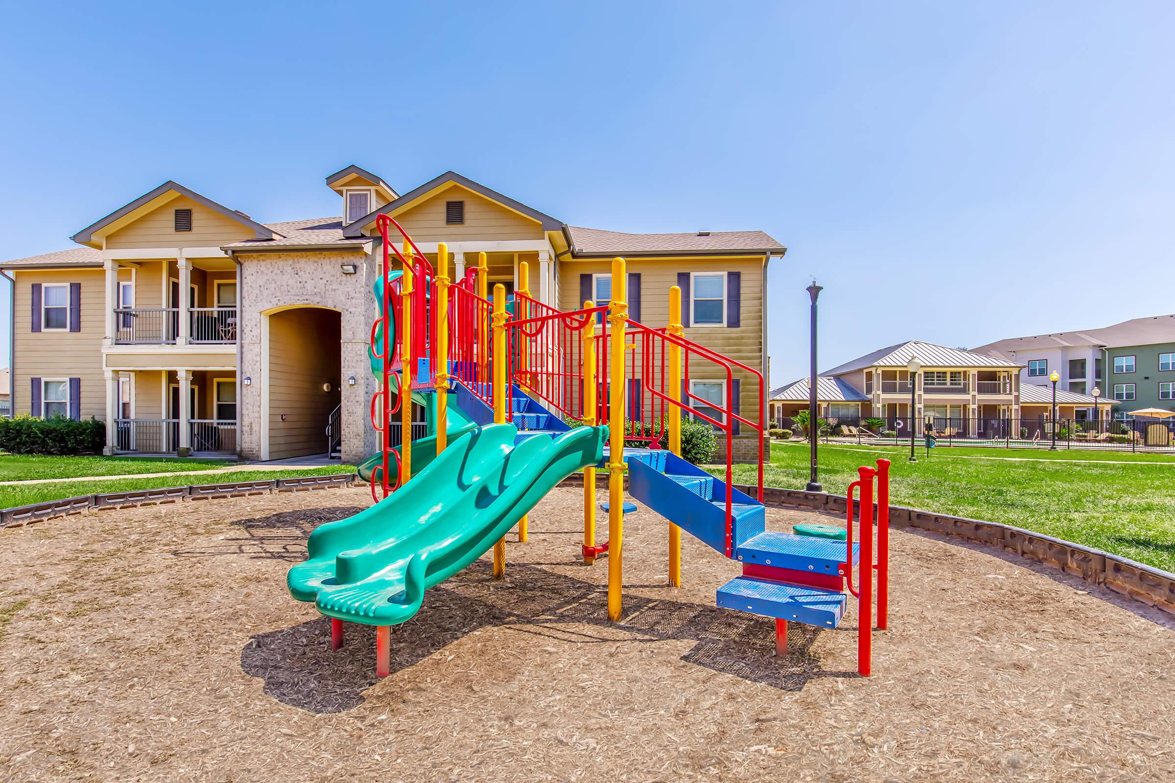 Colorful playground with a green slide and bright red and blue climbing structures, surrounded by mulch, set in front of a residential apartment building. The area is sunny, with additional playground equipment visible in the background and well-maintained green grass.