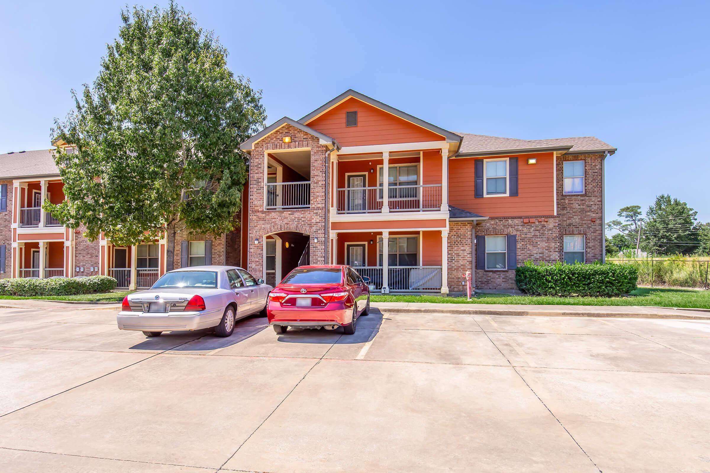 A two-story apartment building with orange and brick exterior. It has balconies on the second floor and a large tree in front. Two parked cars, one silver and one red, are in the foreground, with a well-maintained grassy area and trees in the background under a clear blue sky.