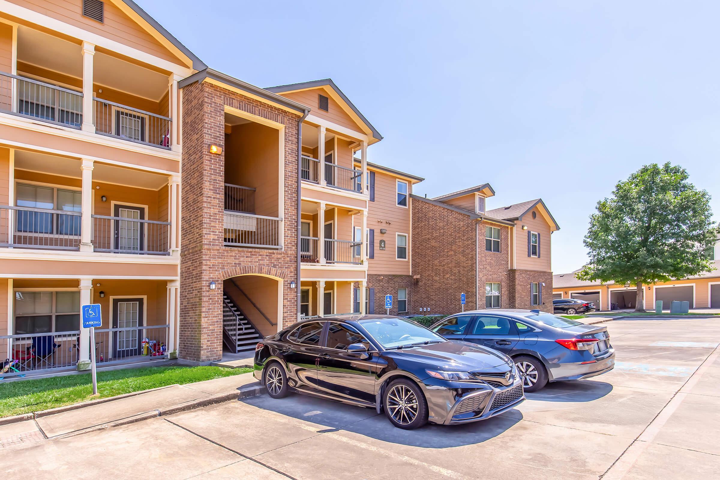 Exterior view of a multi-family apartment building with two levels. A black car is parked in the foreground next to a silver car. The building features a mix of brick and wood siding, with balconies on the upper level and a grassy area nearby. Clear blue sky overhead.