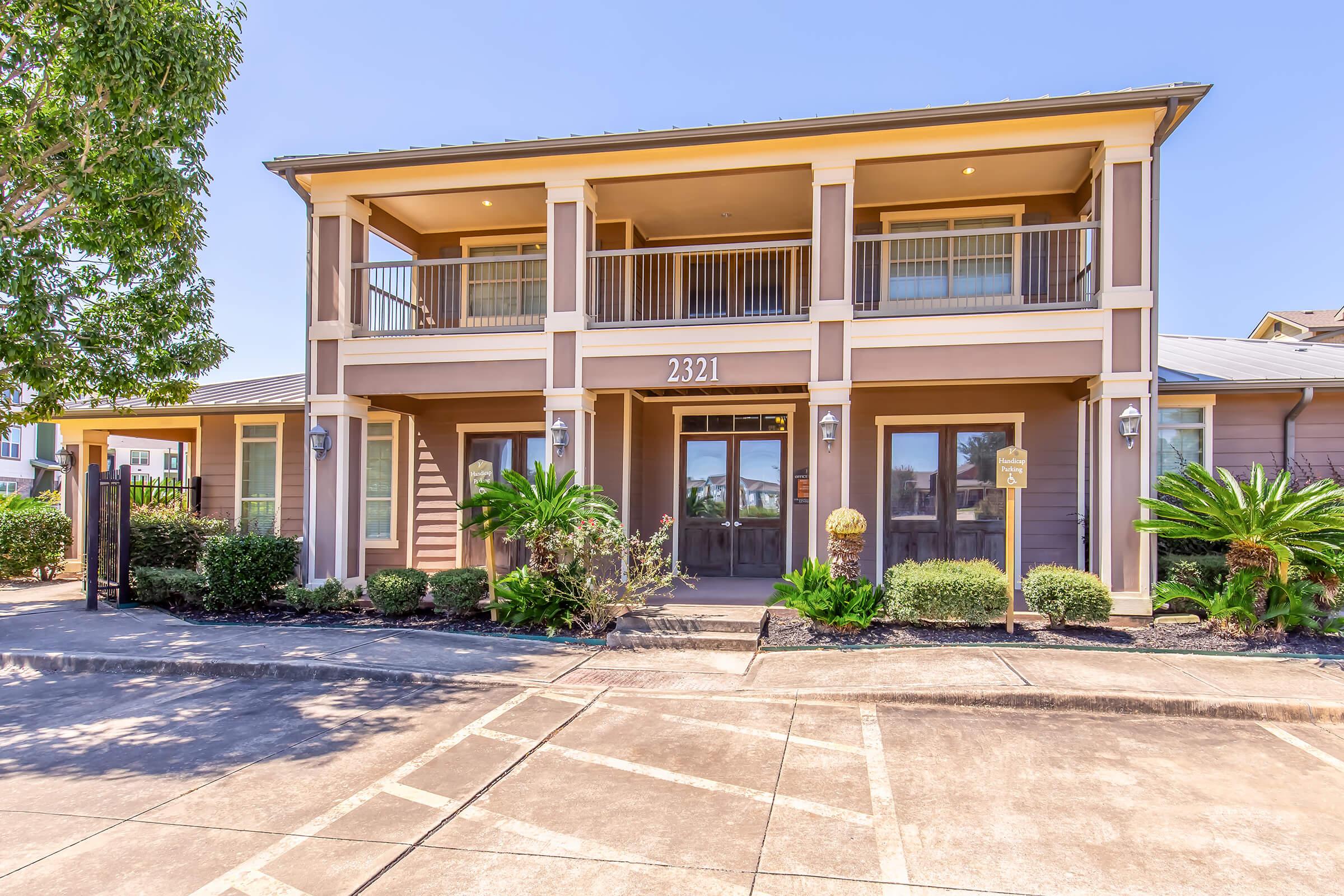 Two-story residential building featuring a welcoming entrance with double doors, large windows, and balconies on the upper floor. Surrounded by neatly trimmed shrubs and small palm trees, the facade has a modern design with light-colored siding. A concrete parking area is visible in front.