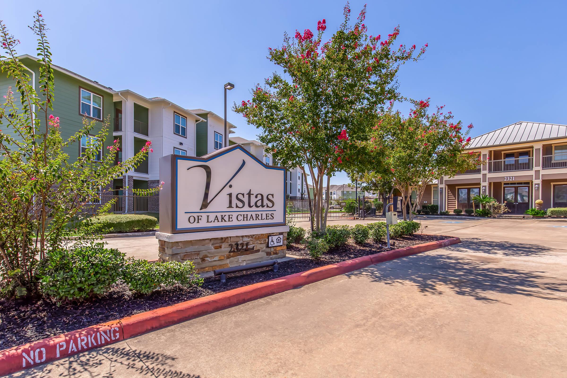 Sign for "Vistas of Lake Charles" prominently displayed at the entrance, surrounded by landscaped greenery and flowering plants. In the background, modern apartment buildings can be seen, indicating a residential complex. Clear blue sky overhead enhances the inviting atmosphere.