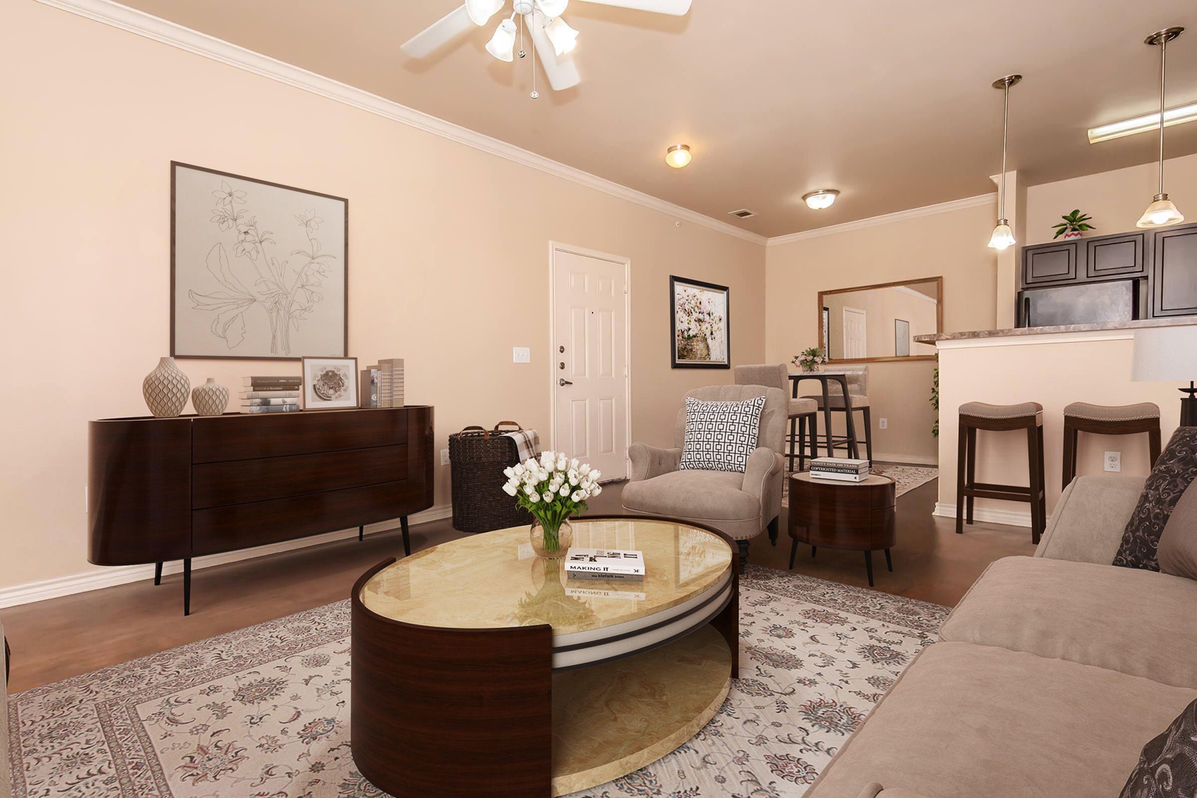 A cozy living room featuring a light-colored wall and ceiling fan, a circular coffee table with decorative items, a plush sofa, and a stylish side chair. The space includes a wooden sideboard, a potted flower arrangement, and a patterned area rug, creating a warm and inviting atmosphere.