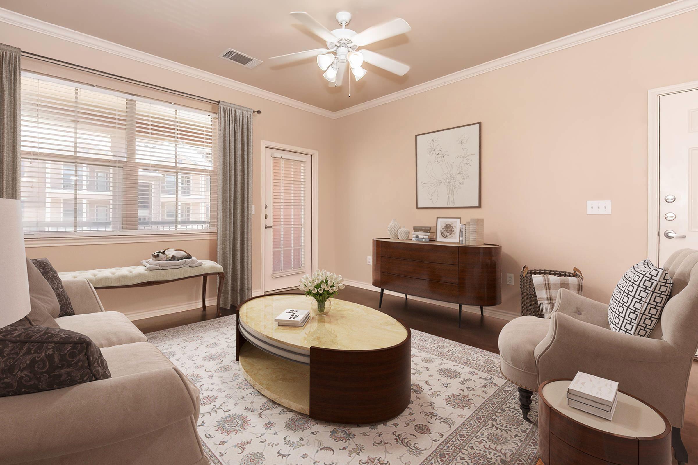 A cozy living room with light-colored walls and a ceiling fan. It features a beige sofa, a patterned area rug, and a round coffee table with a gold top. A wooden console table holds decorative items, and large windows with curtains let in natural light. A door leads to an outdoor space.