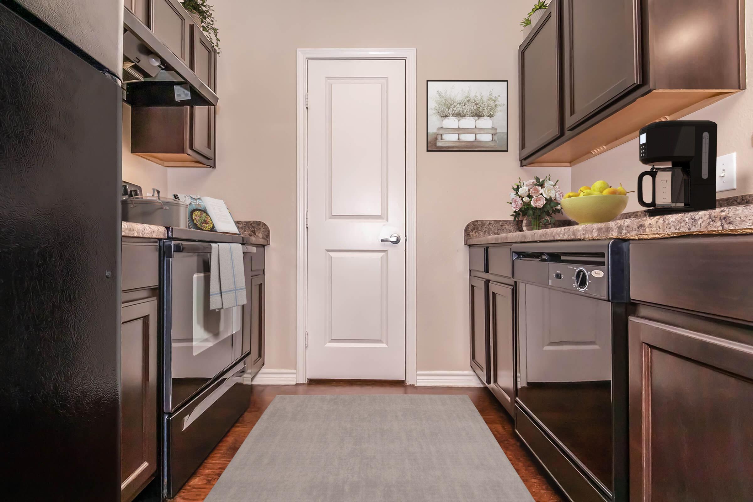 A modern kitchen with dark wooden cabinets, stainless steel appliances, and a gray area rug. The countertop features a bowl of fruit and a flower arrangement. A white door leads to another room, and a framed picture hangs on the wall, enhancing the cozy atmosphere.