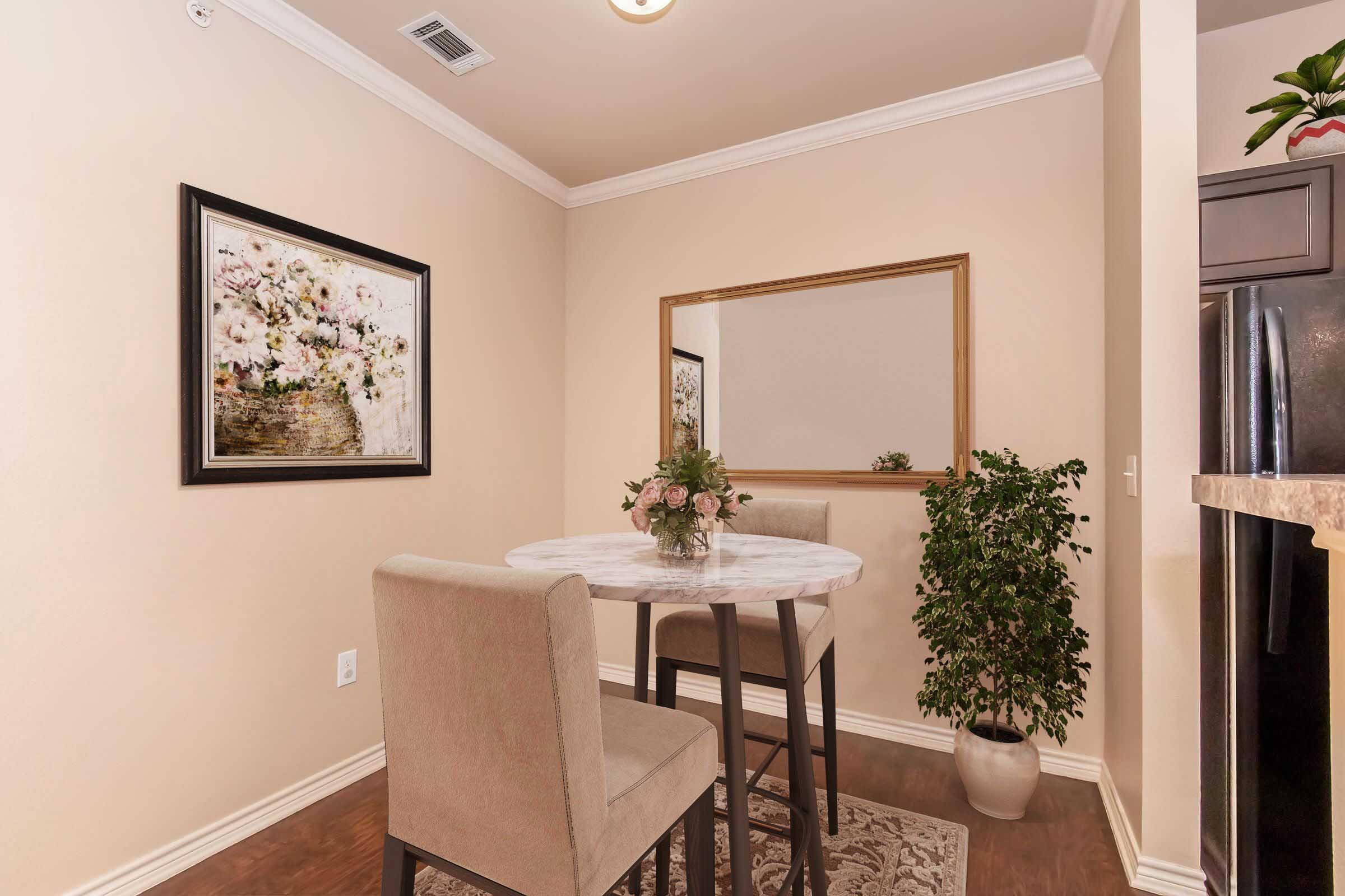 A small dining area featuring a round marble table surrounded by two beige chairs. There's a floral painting on the wall, a large mirror reflecting the space, and a potted plant in the corner. The walls are painted a warm beige, creating a cozy atmosphere.