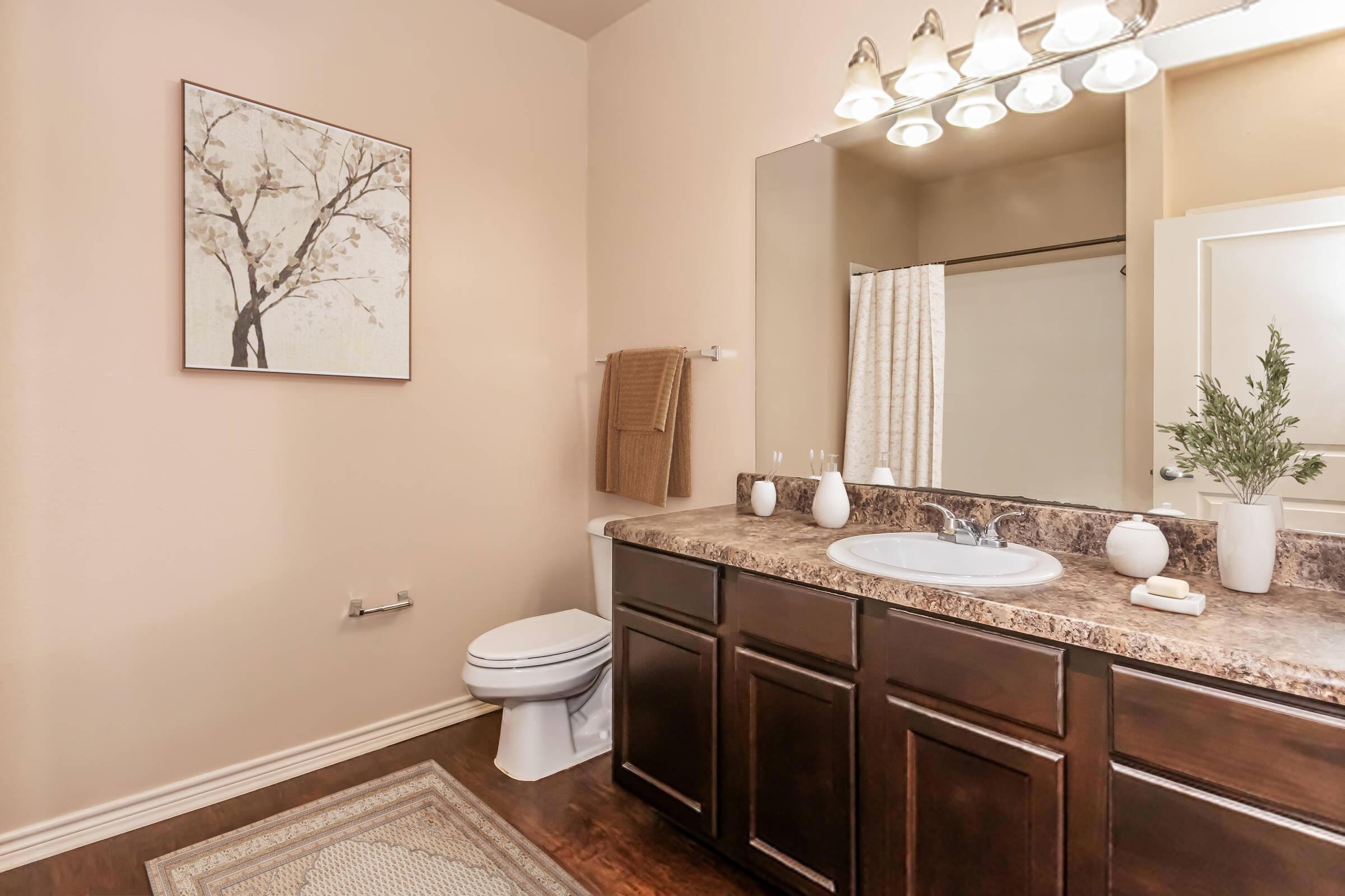 A well-lit bathroom featuring a dark wood vanity with a sink, a large mirror above it, and a decorative tree painting on the wall. There are towels hanging nearby, a shower curtain in the background, and a small plant on the counter. The floor is covered with a light-colored rug.