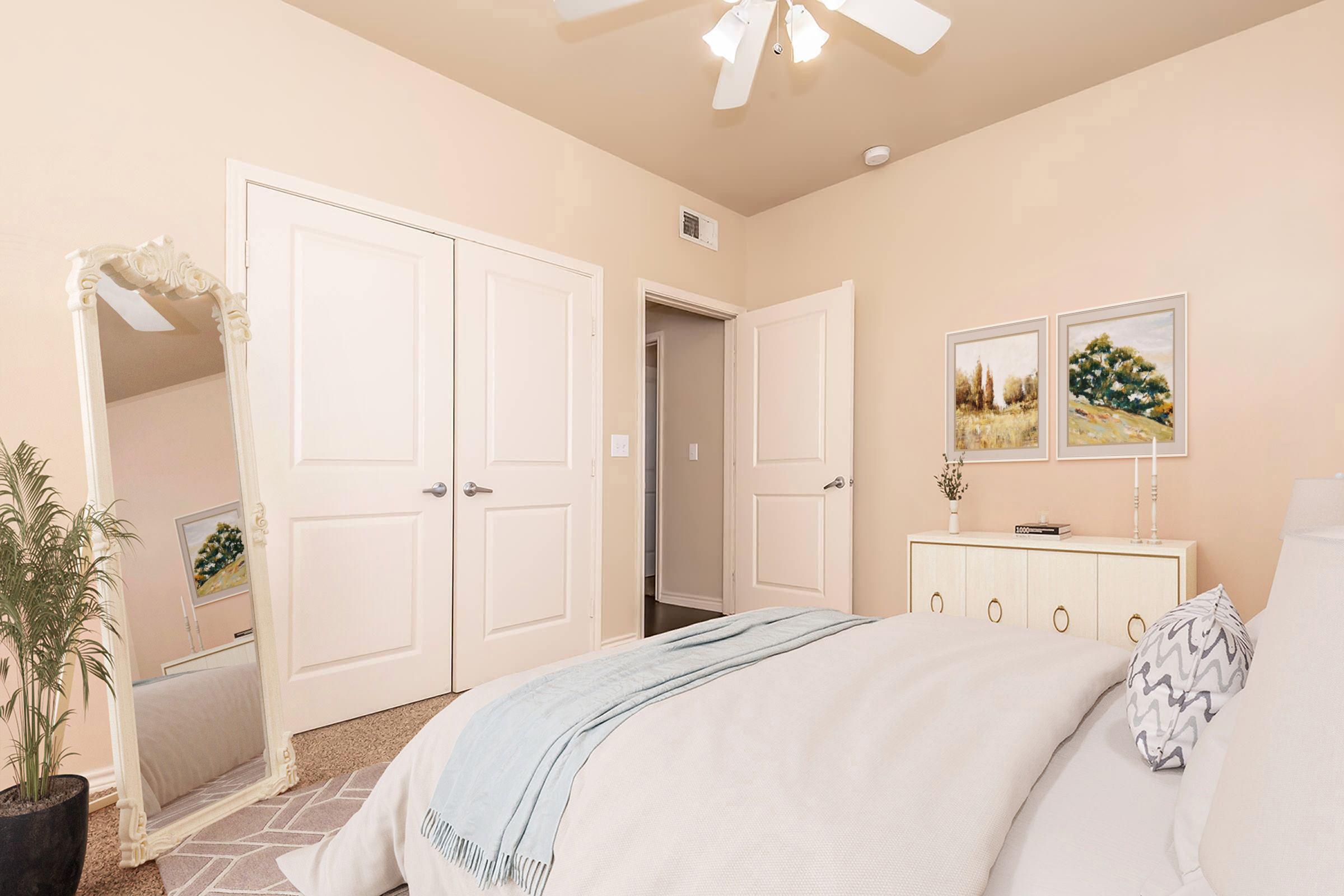 A cozy bedroom featuring a light-colored wall, a neatly made bed with a soft blanket, and decorative pillows. A tall mirror reflects the room, and two wooden doors lead to another space. On the wall, two framed artworks depict serene landscapes, adding a touch of elegance to the ambiance. A small plant sits next to the dresser.